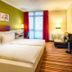 Leonardo Hotel & Residenz München guest room with twin beds, sofa, TV, and window with red curtains