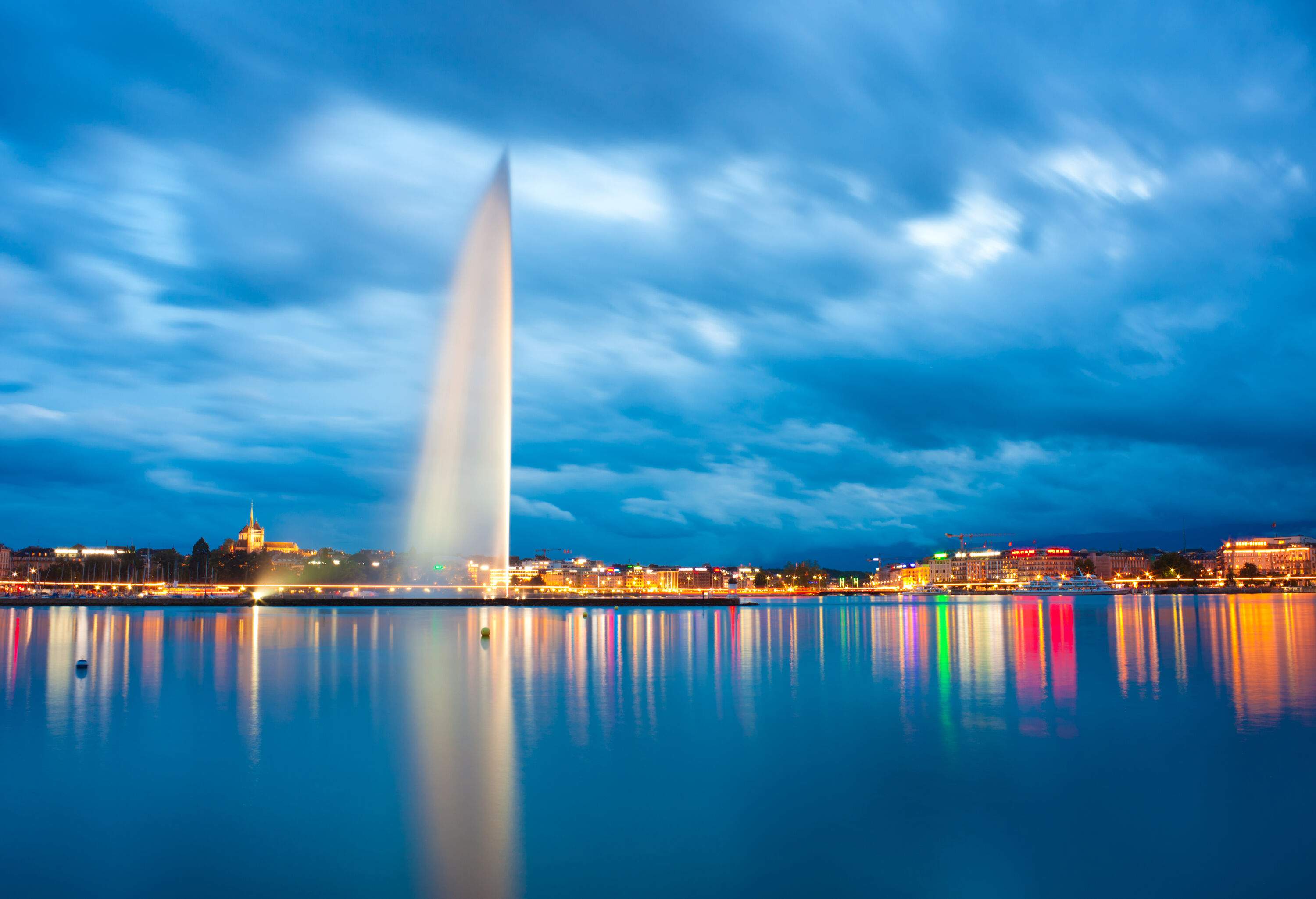 The Jet d'Eau jutting into the sky as streaks of night lights reflect on the lake.