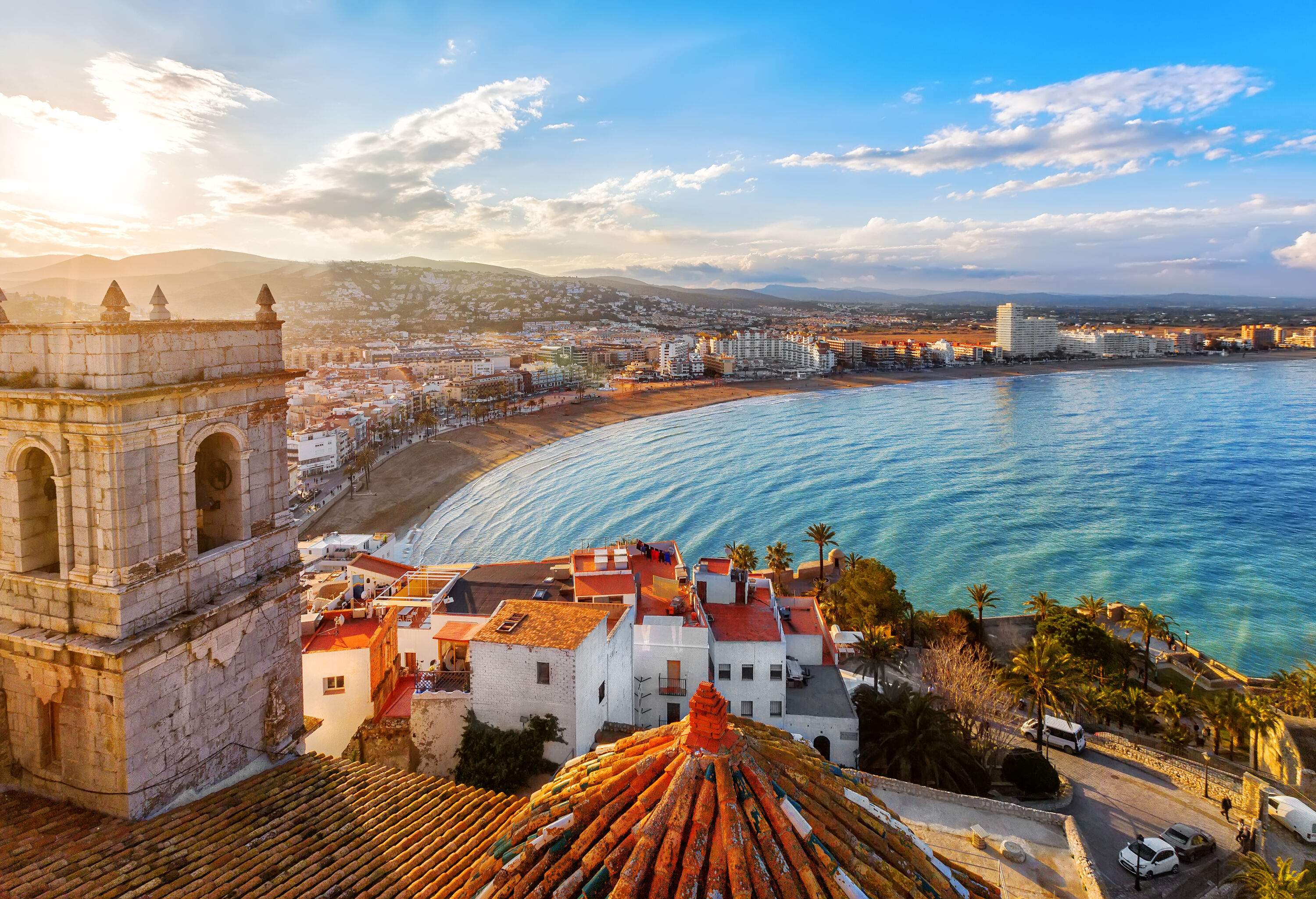 On top of a castle's roof is a view of a tightly packed city along the sea.