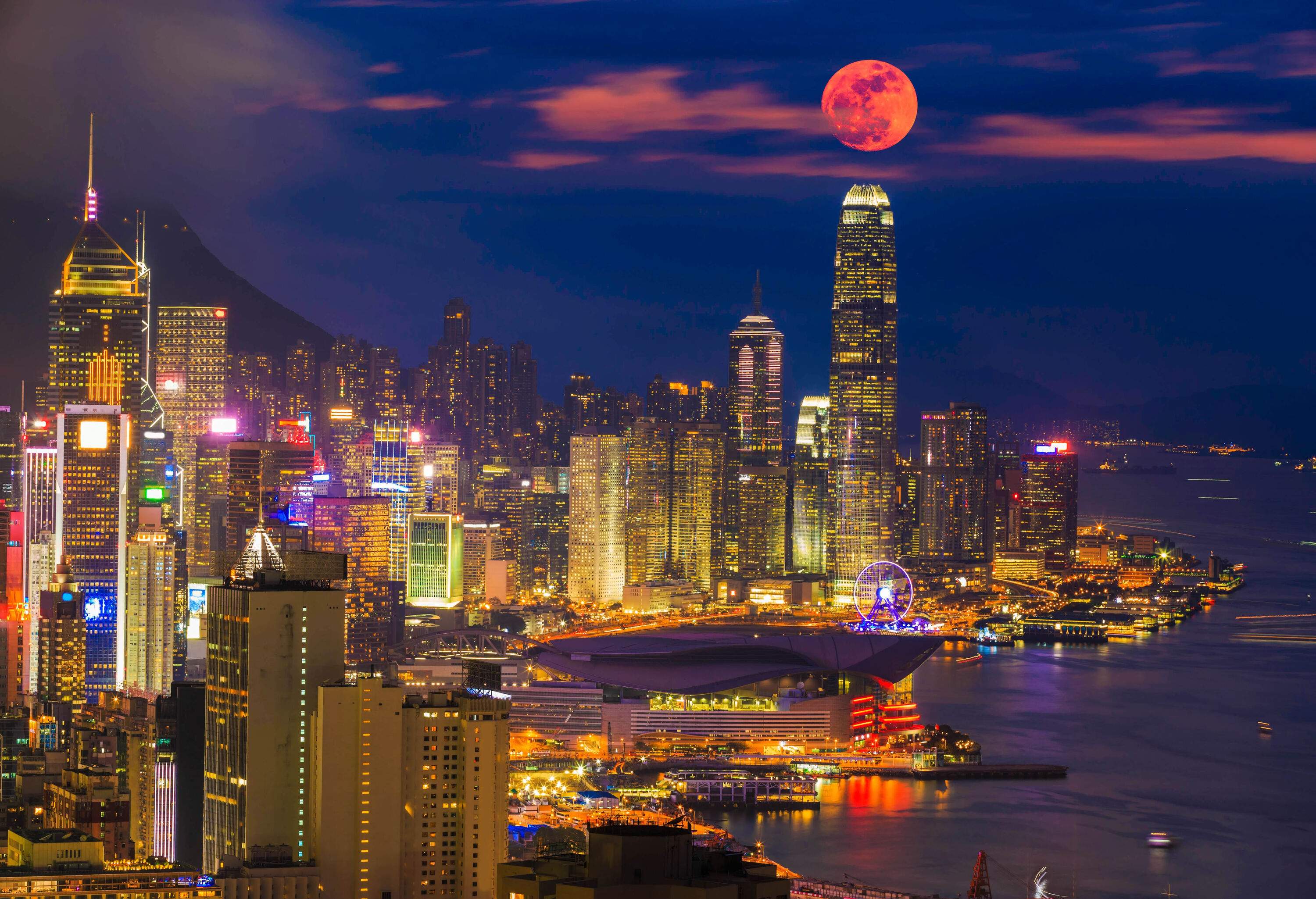 A bustling coastal city sparkles under the glow of bright skyscrapers, the scene is further illuminated by a radiant full moon in the sky.