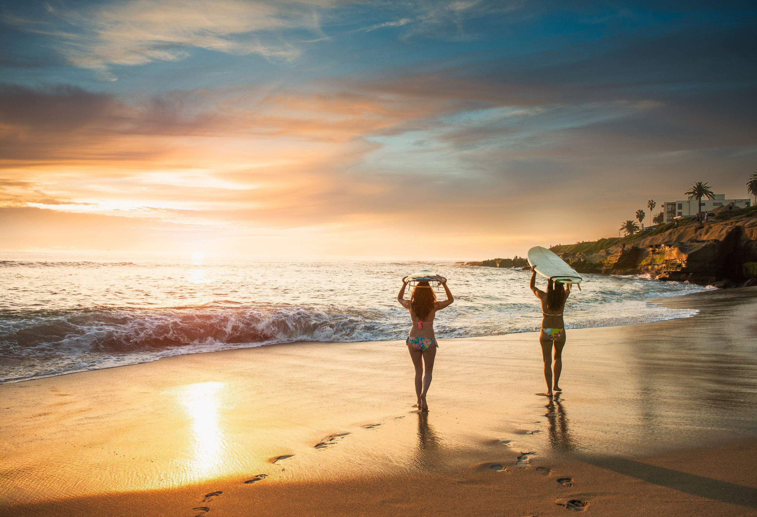 Rear view of two ladies in colourful bikinis carrying surfboards over their heads while walking on the shore against the dramatic sky.