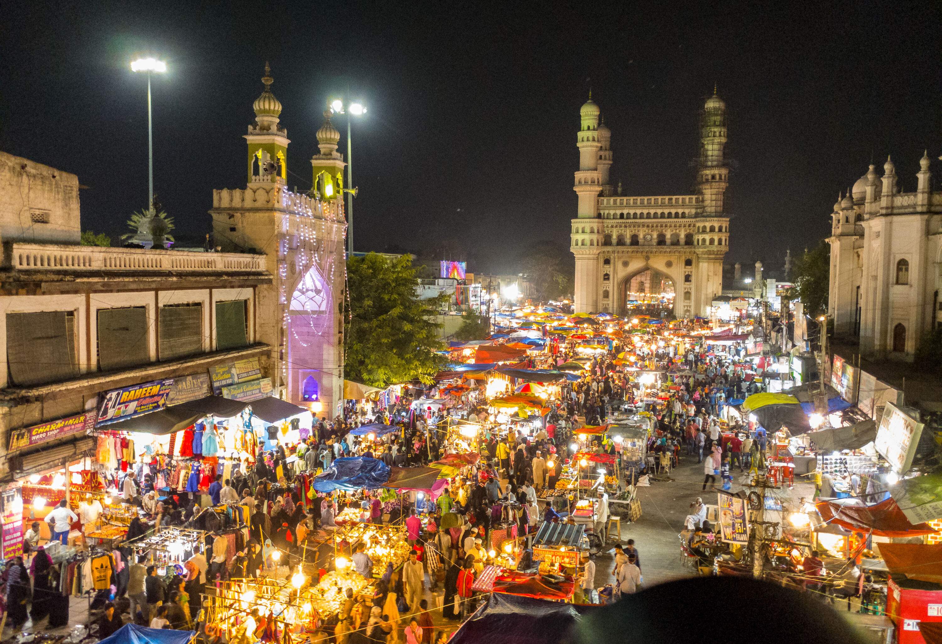 A bustling crowd fills the lively street adorned with shops and buildings, while a prominent gateway stands in the background.