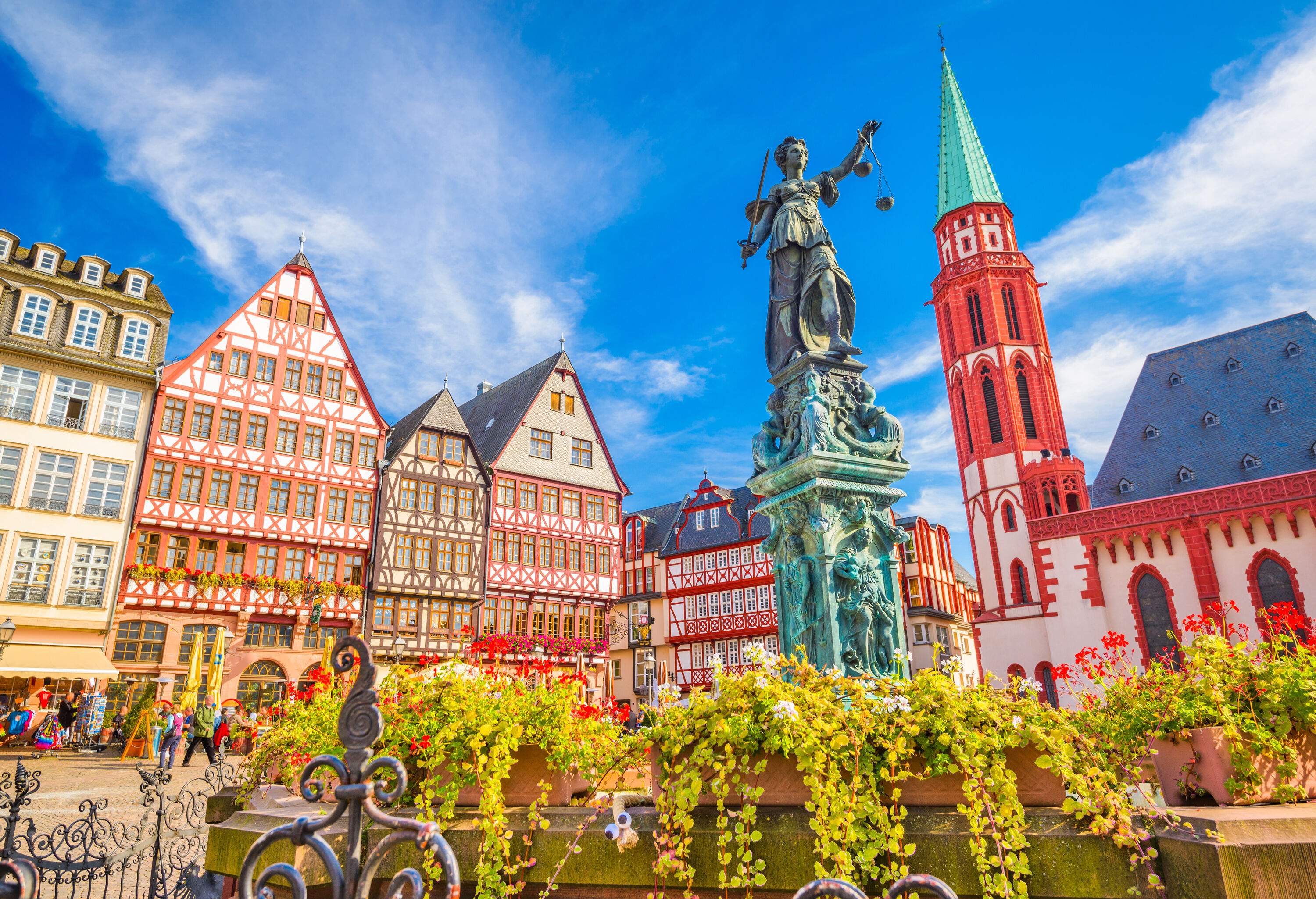 A cobbled town square with a statue in the centre surrounded by half-timbered ornate and colourful townhouses and a small brick gothic church with a towering spire.
