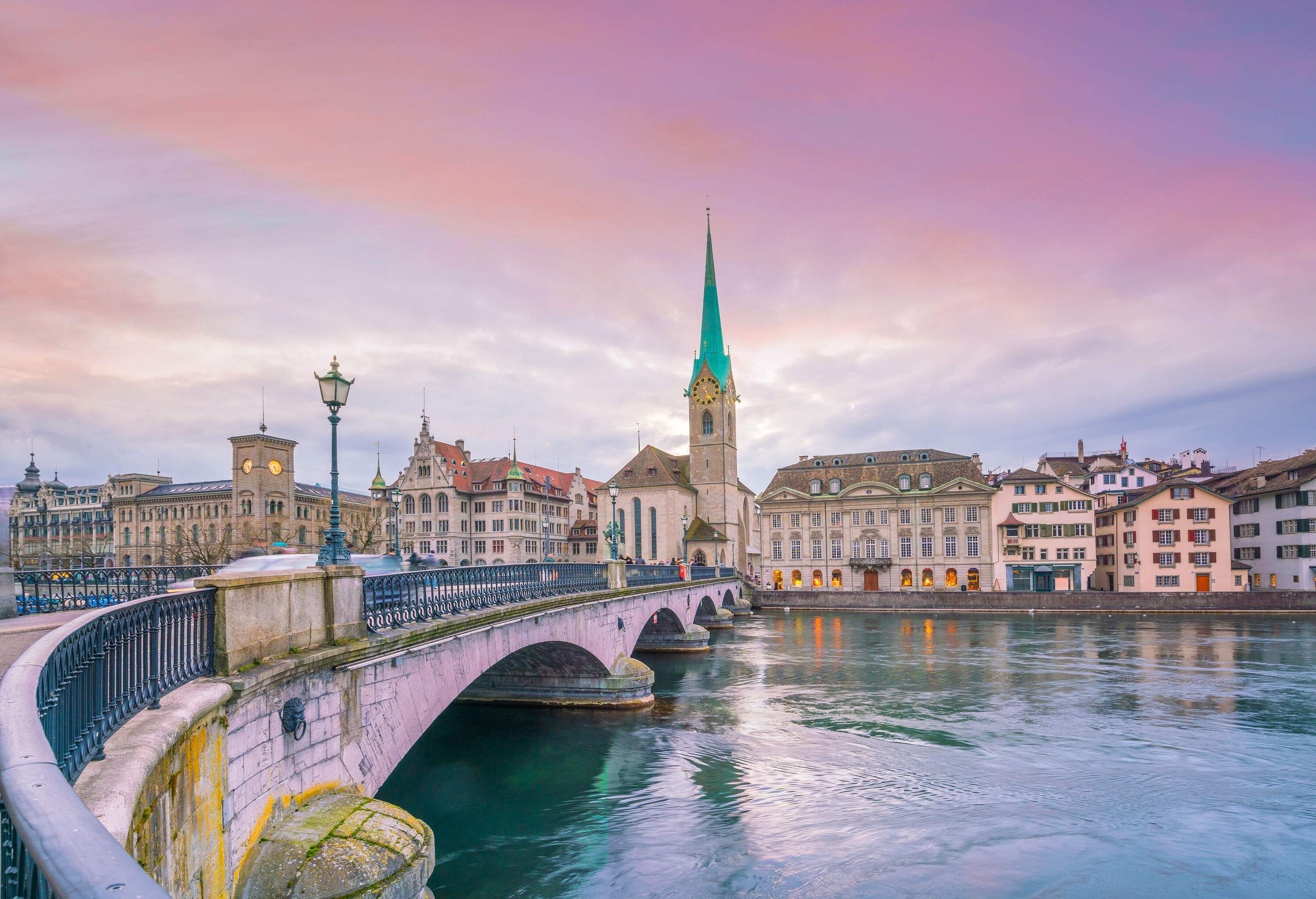 A pointed clock tower rises above the cityscape as seen from a bridge across a calm river.