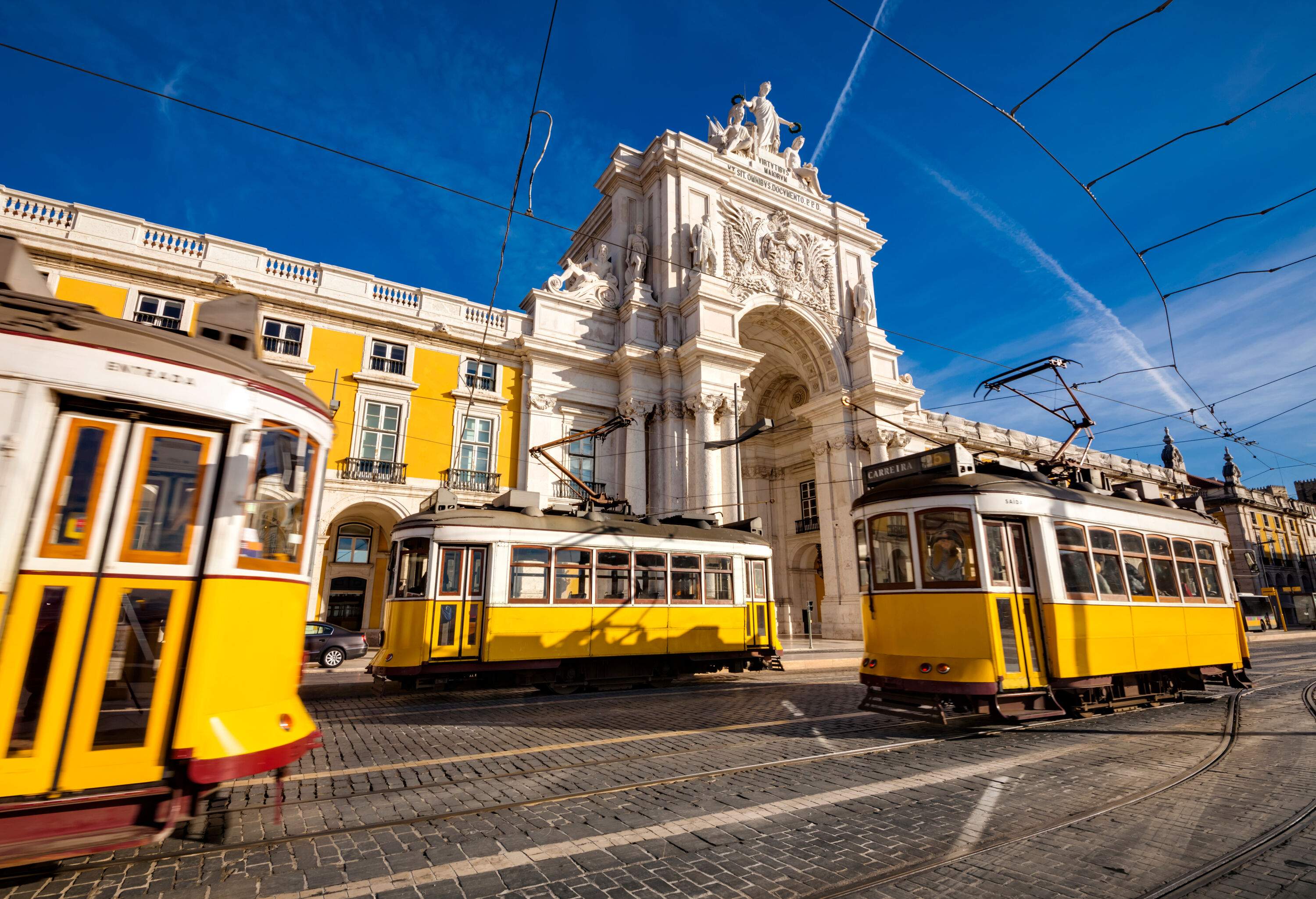 Three yellow trams travel along a yellow building with a pillared memorial arch in the middle.