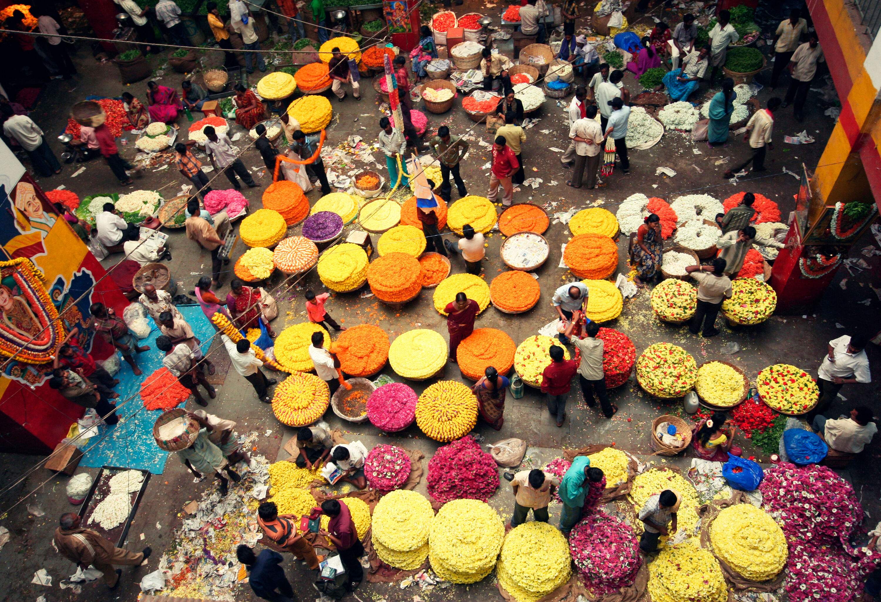 A crowded market with vendors selling vibrant red and orange flower garlands in huge baskets.