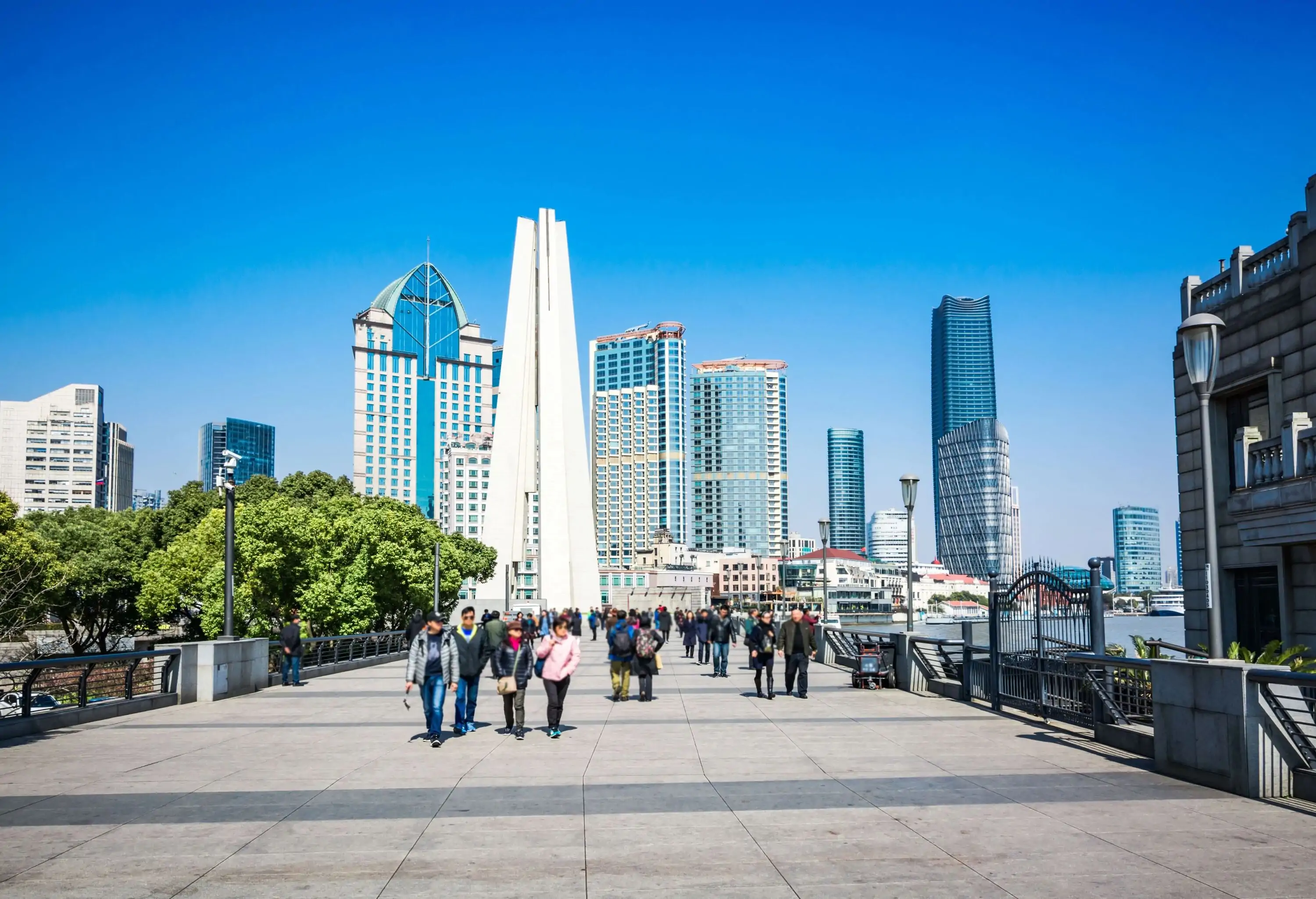 A wide, bustling walkway, enlivened by the rhythmic footsteps of people strolling, gracefully leads the way towards a majestic cluster of towering buildings, forming a captivating urban corridor.