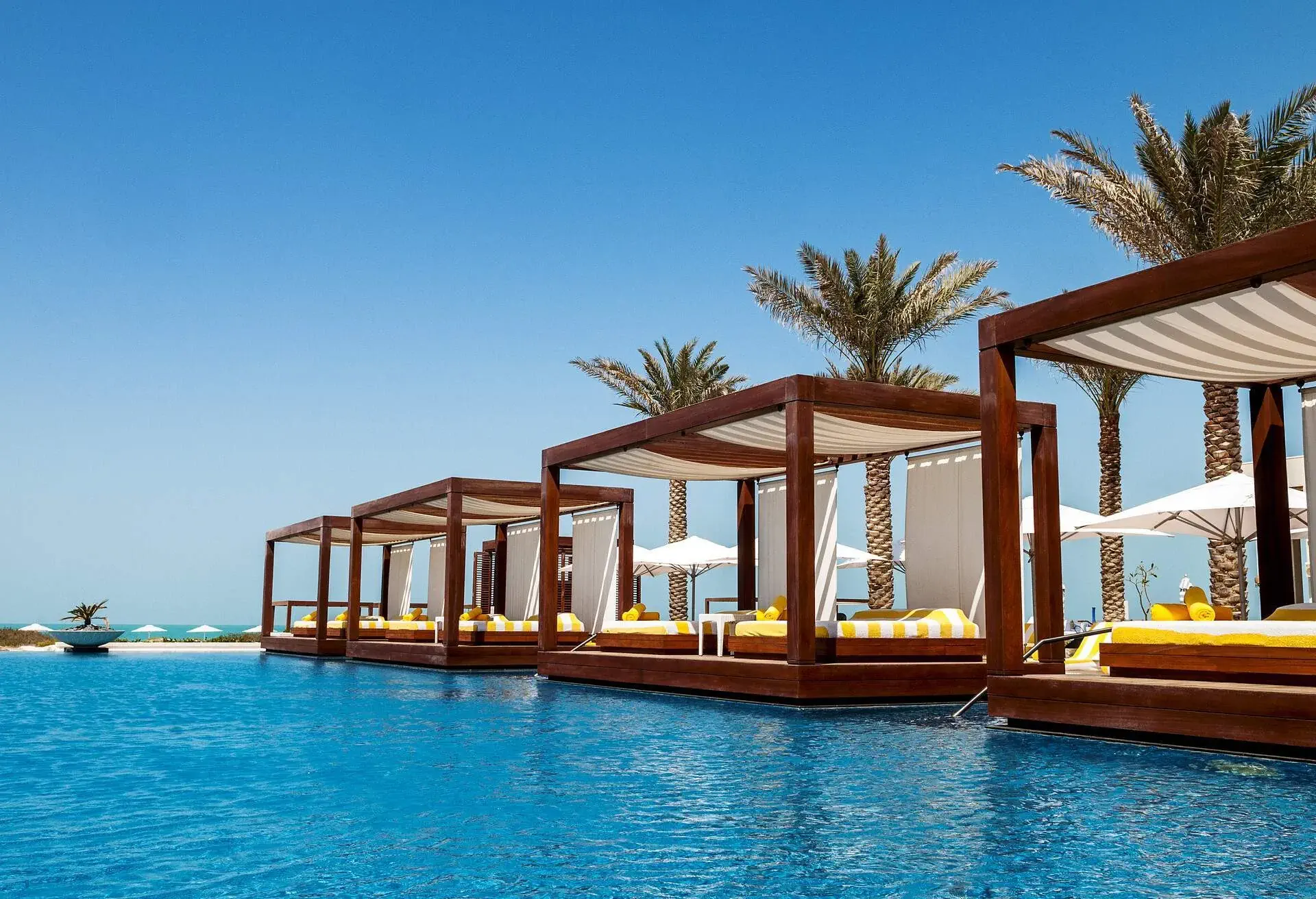View of sunbeds by the pool, near to a white sand beach and with some palm trees in the background