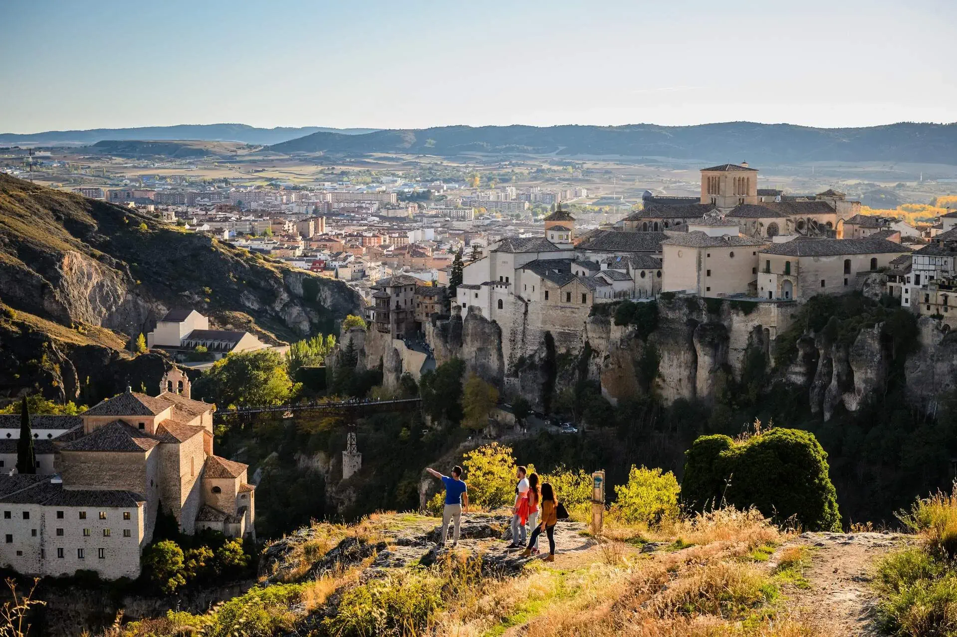 Several tourists are atop the hill with a view of hanging houses on the cliff and the cityscape in the valley in the distance.