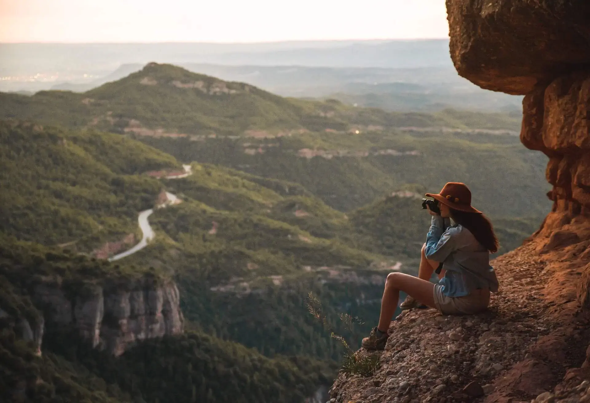 A woman sits on a cliff's edge while taking photos of a hillside road on a mountainous terrain.