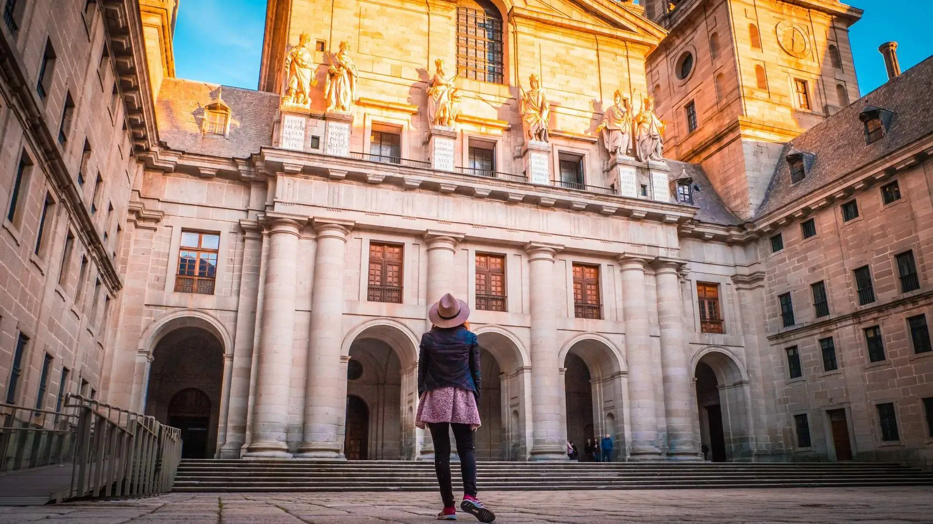 A woman stands in front of a basilica with massive white columns and arched entrances, looking up at the sun-lit human sculptures.