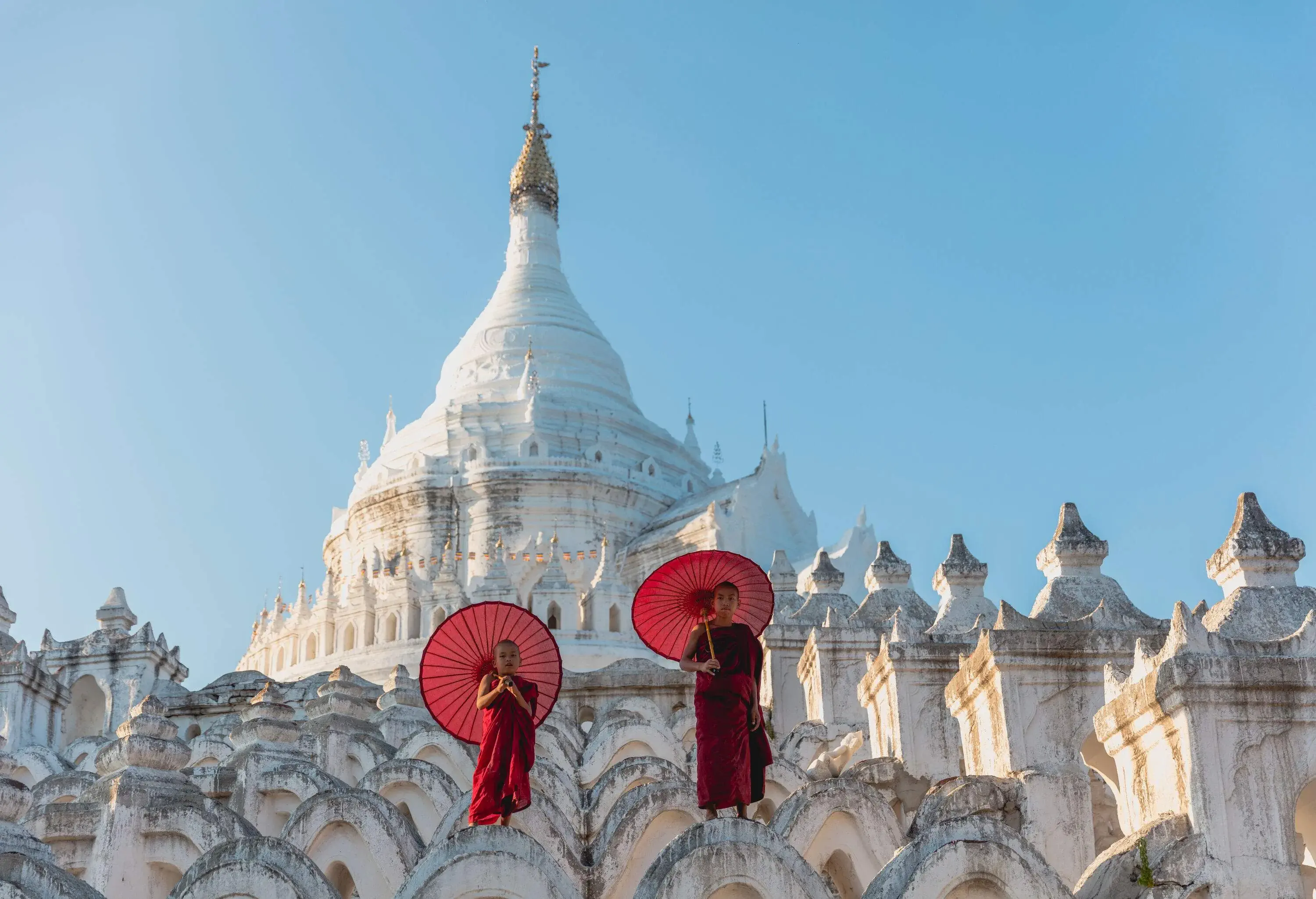 Two young Buddhist novices with red parasols stand on the wavy layers of a large white pagoda.
