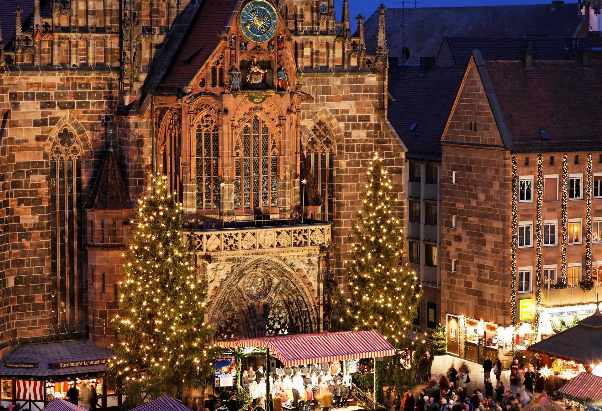 An elaborately decorated brick cathedral in Gothic style with tall Christmas trees on either side of its entryway and Christmas shops on the main square.