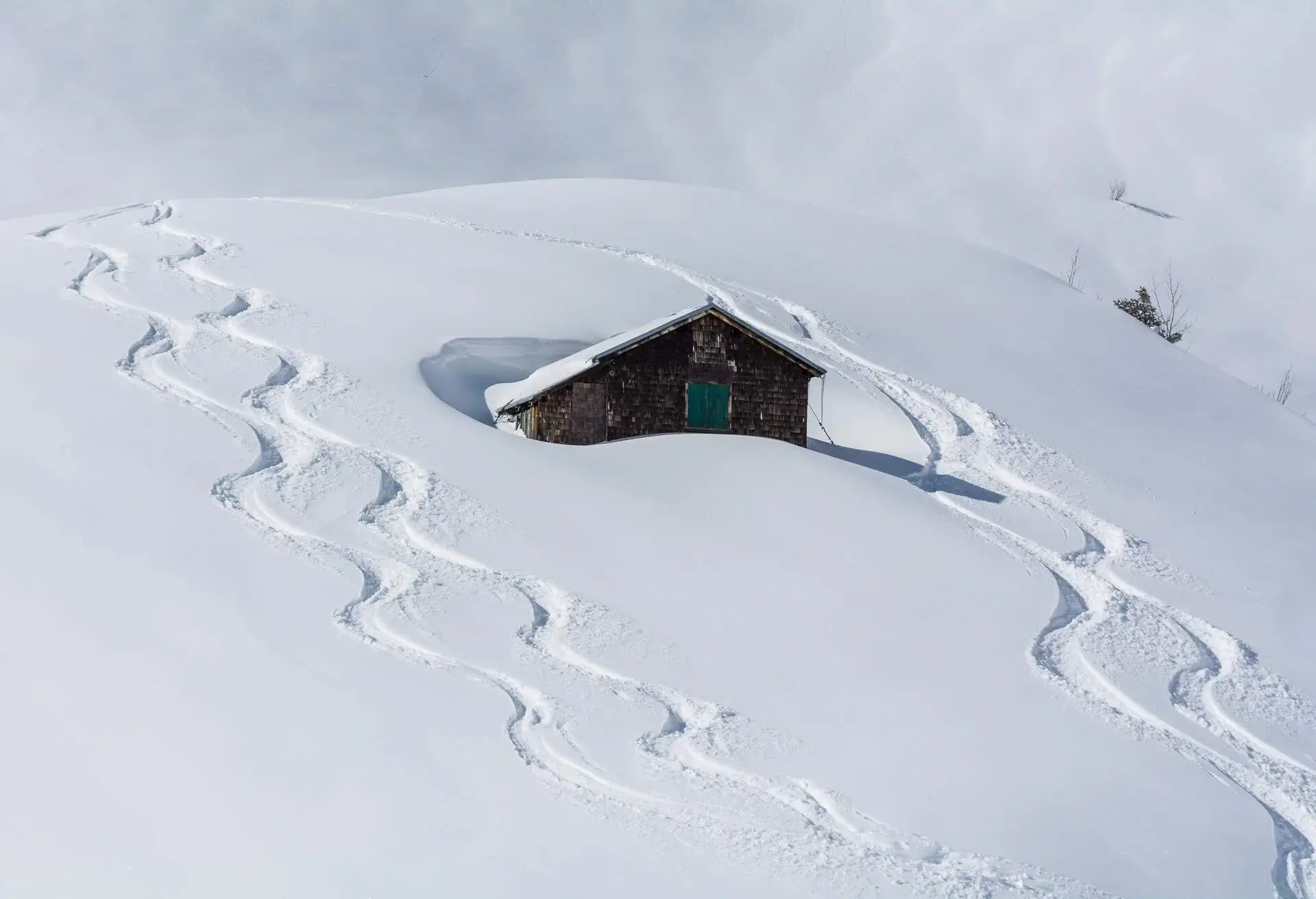 A hillside cabin buried in deep snow with ski tracks.