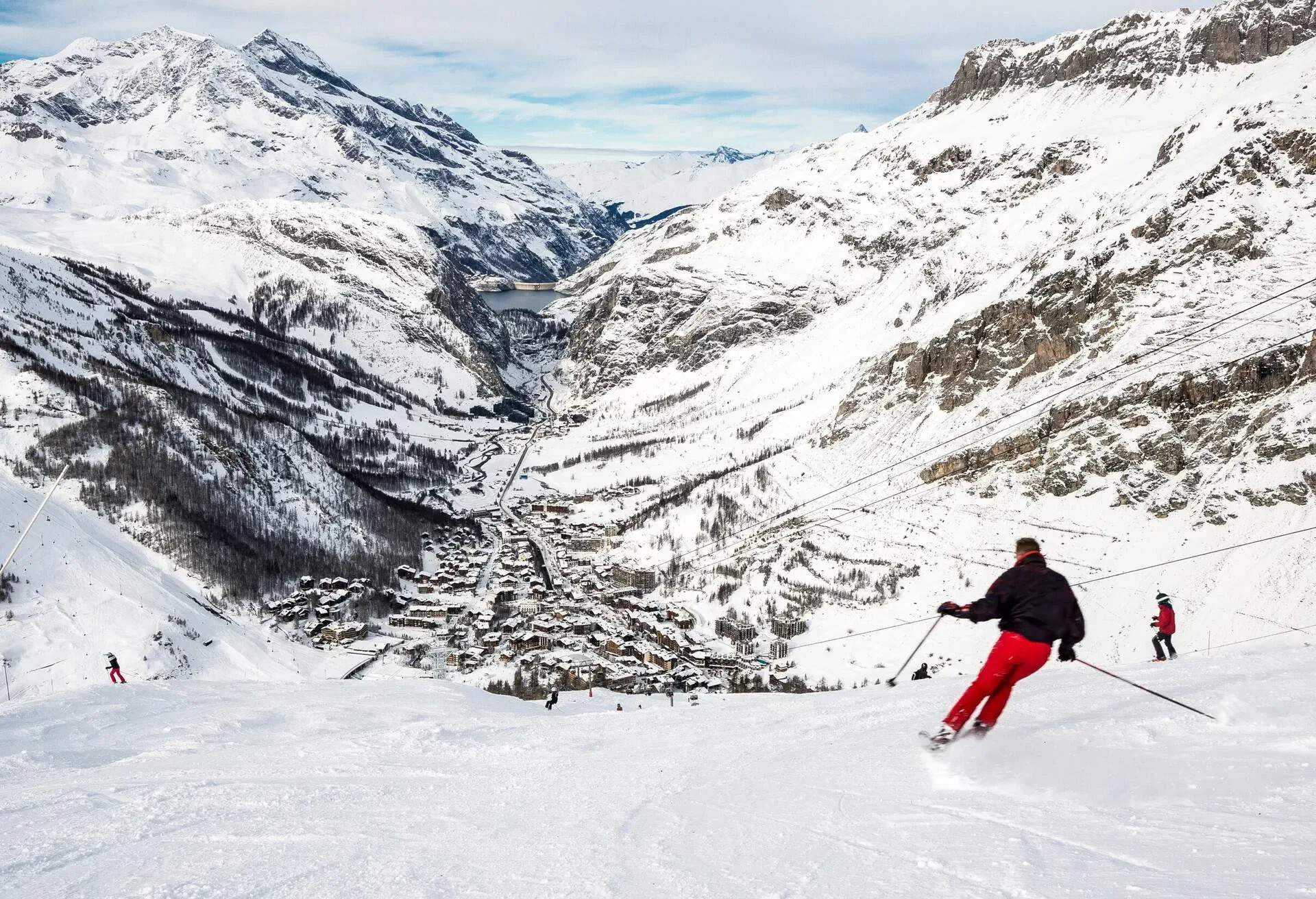 Skiers on a piste overlooking a small snow-covered village at the base of the mountains.