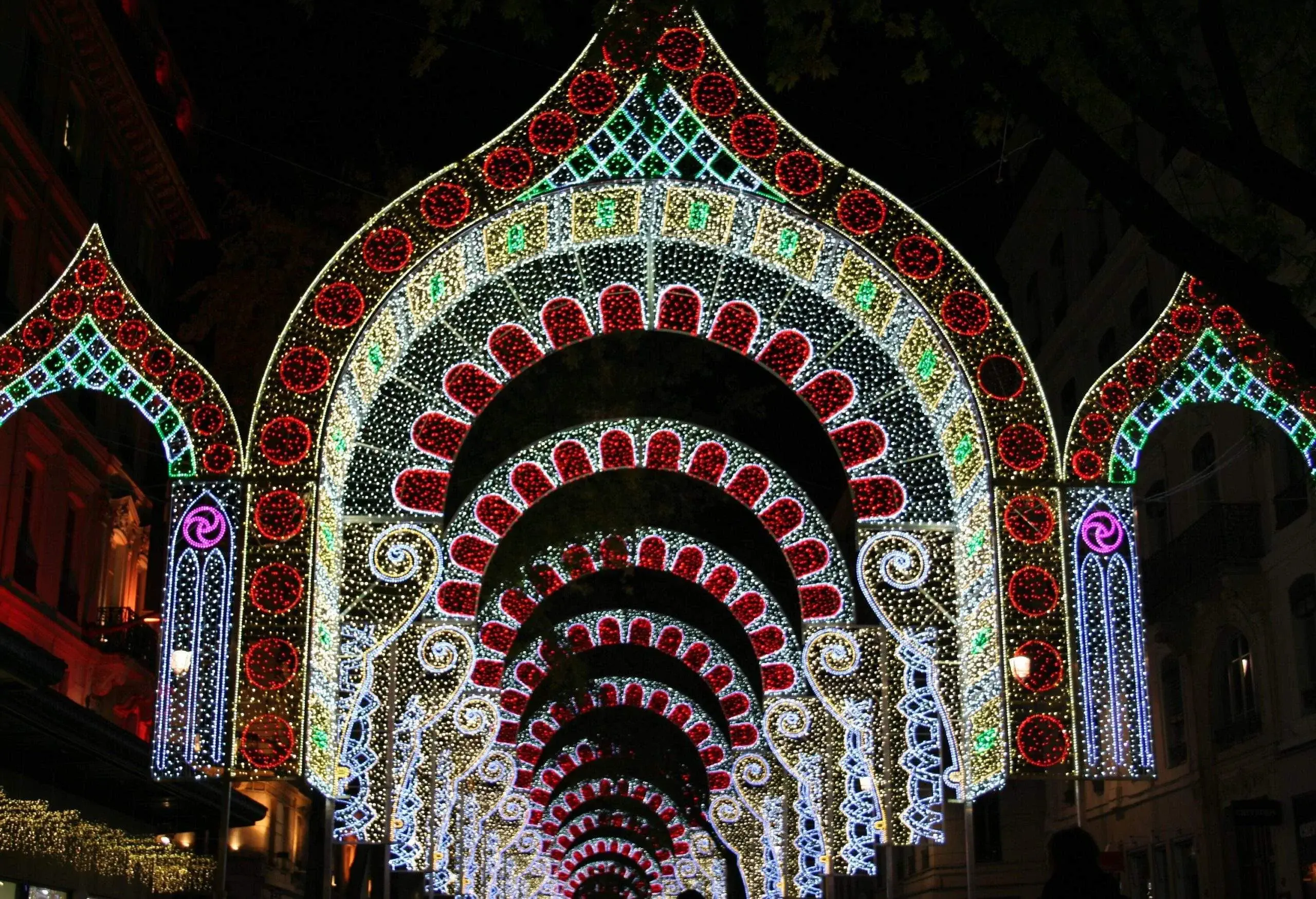 A series of ornate archways illuminated with intricate light patterns in red, white, and green. The arches form a tunnel-like structure, creating a vibrant and festive atmosphere