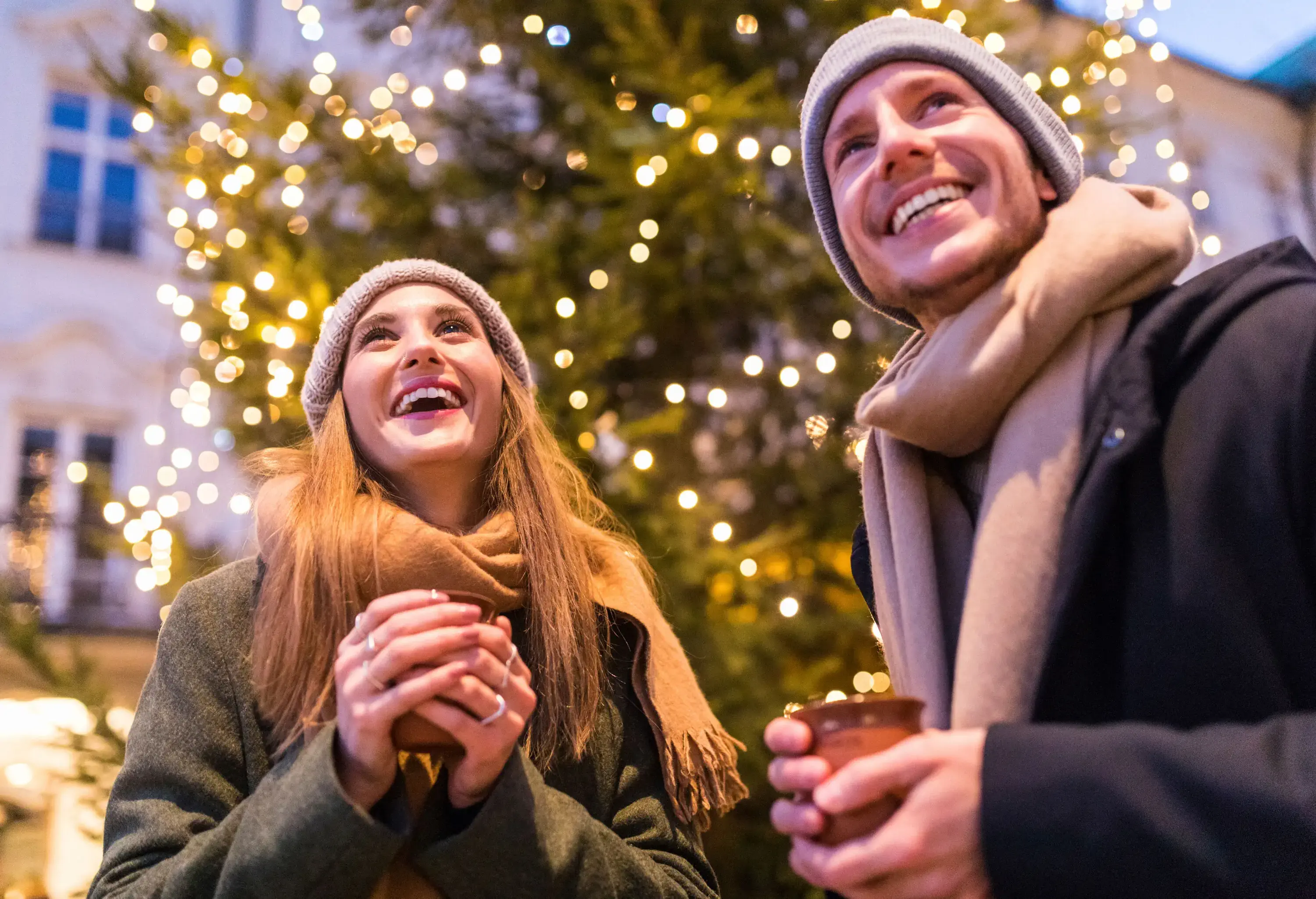 A man and a woman in cosy winter outfits holding warm drinks with a well-lit Christmas tree in the background.