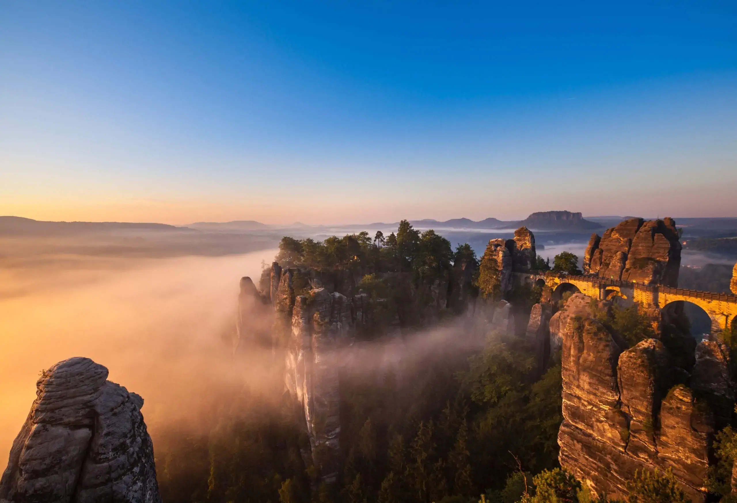 A bridge across a rugged mountain ridge towards the summit of eroded rock formations covered in fog.