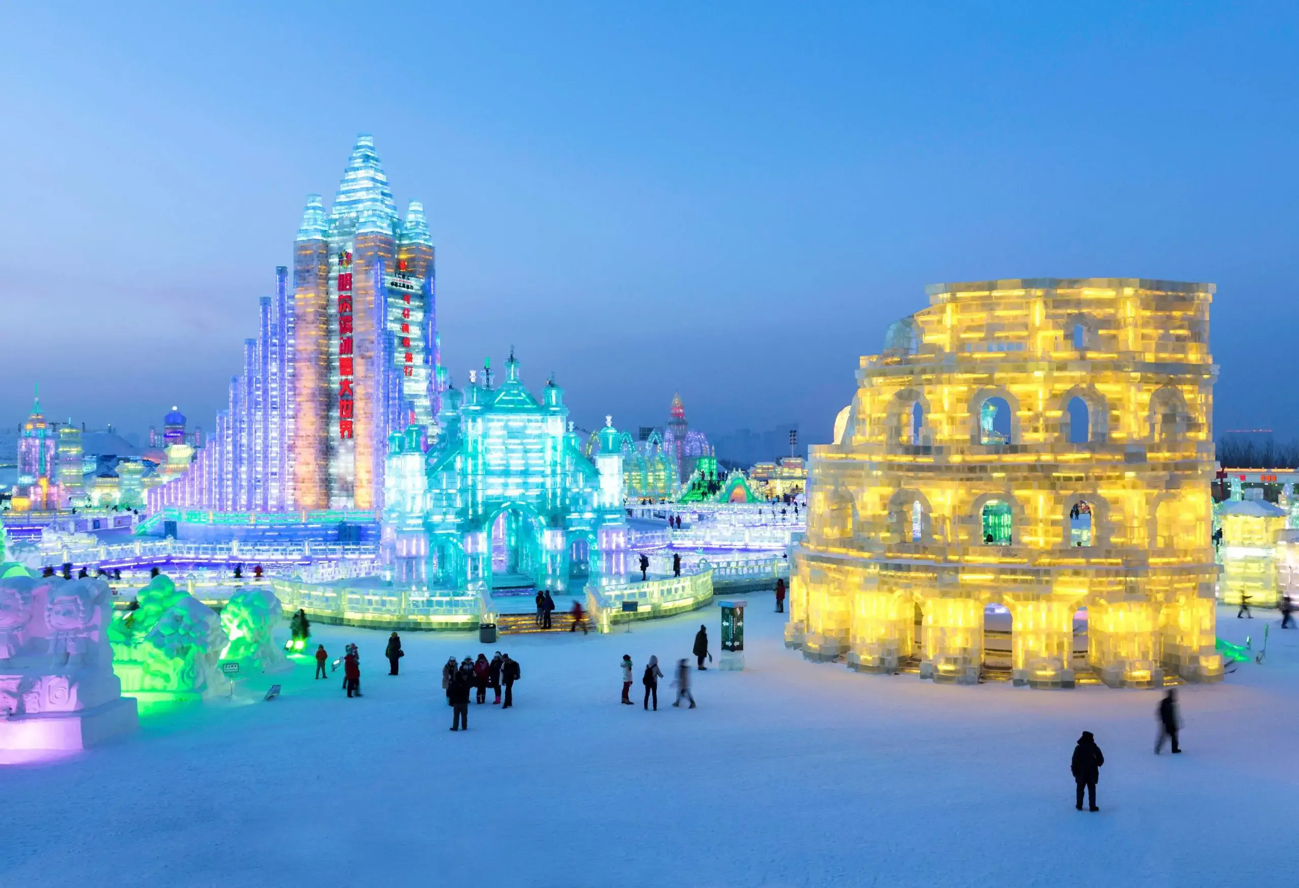 A brightly lit winter ice festival featuring large, intricately designed ice sculptures illuminated with colorful lights. The sculptures include a glowing replica of the Colosseum on the right and tall, ornate structures resembling castles and skyscrapers in the center and left. Visitors are walking and standing on the snow-covered ground, admiring the dazzling displays against a clear evening sky.