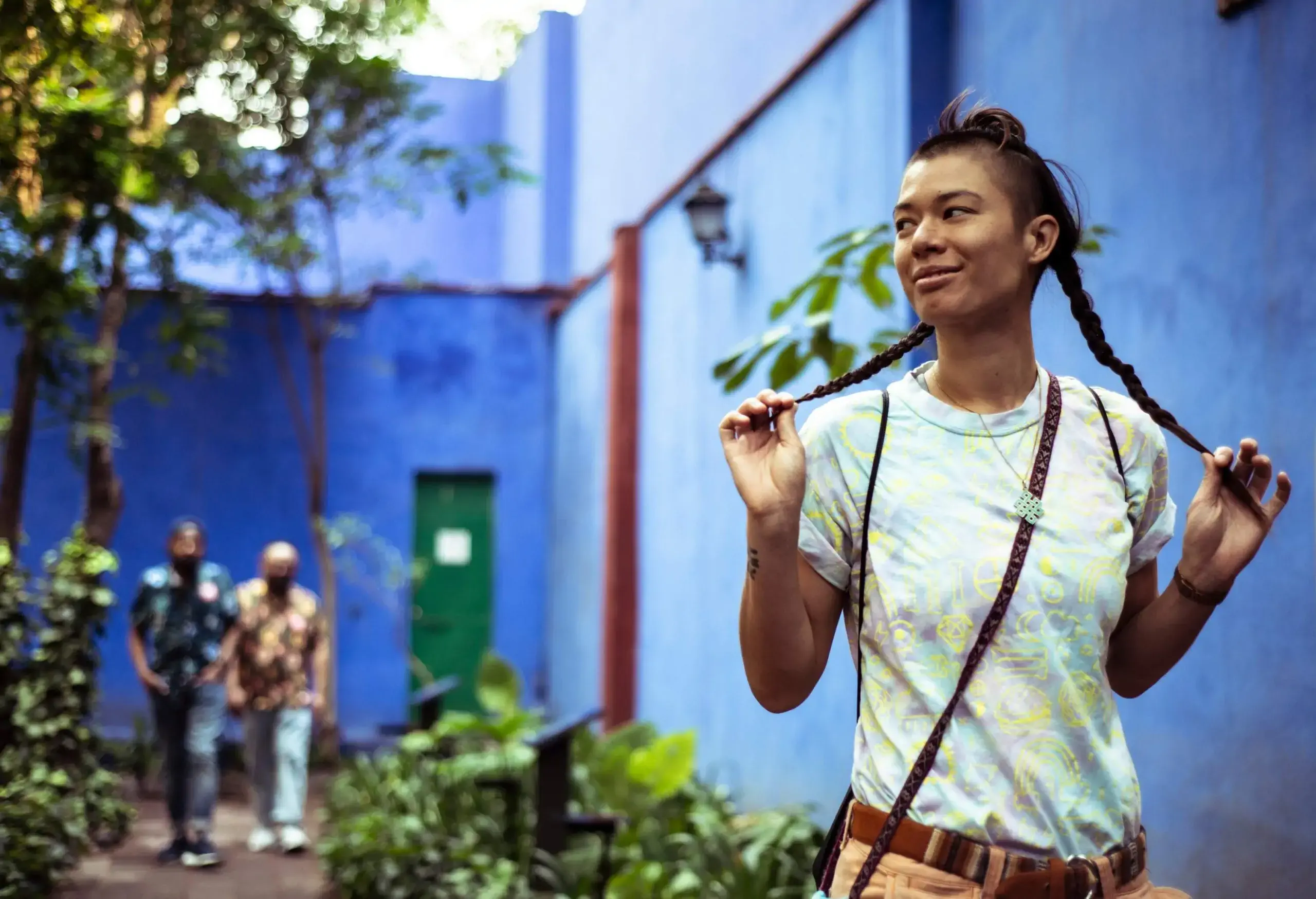 A young woman standing in the yard of a blue-walled building holding her braided hair, with two men walking on a path behind her.