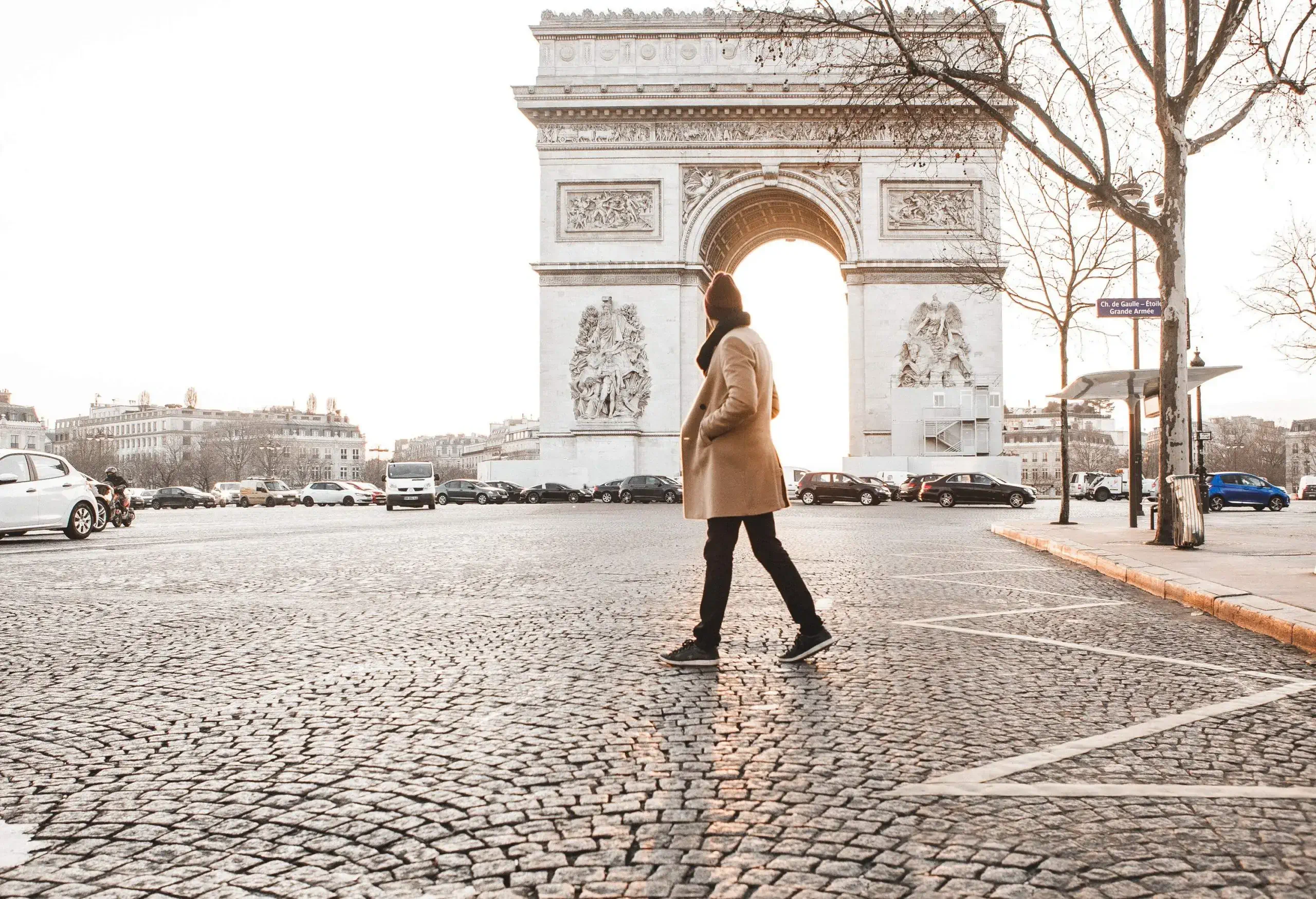A man in a winter coat walking on the cobblestone street in front of the Arc de Triomphe.