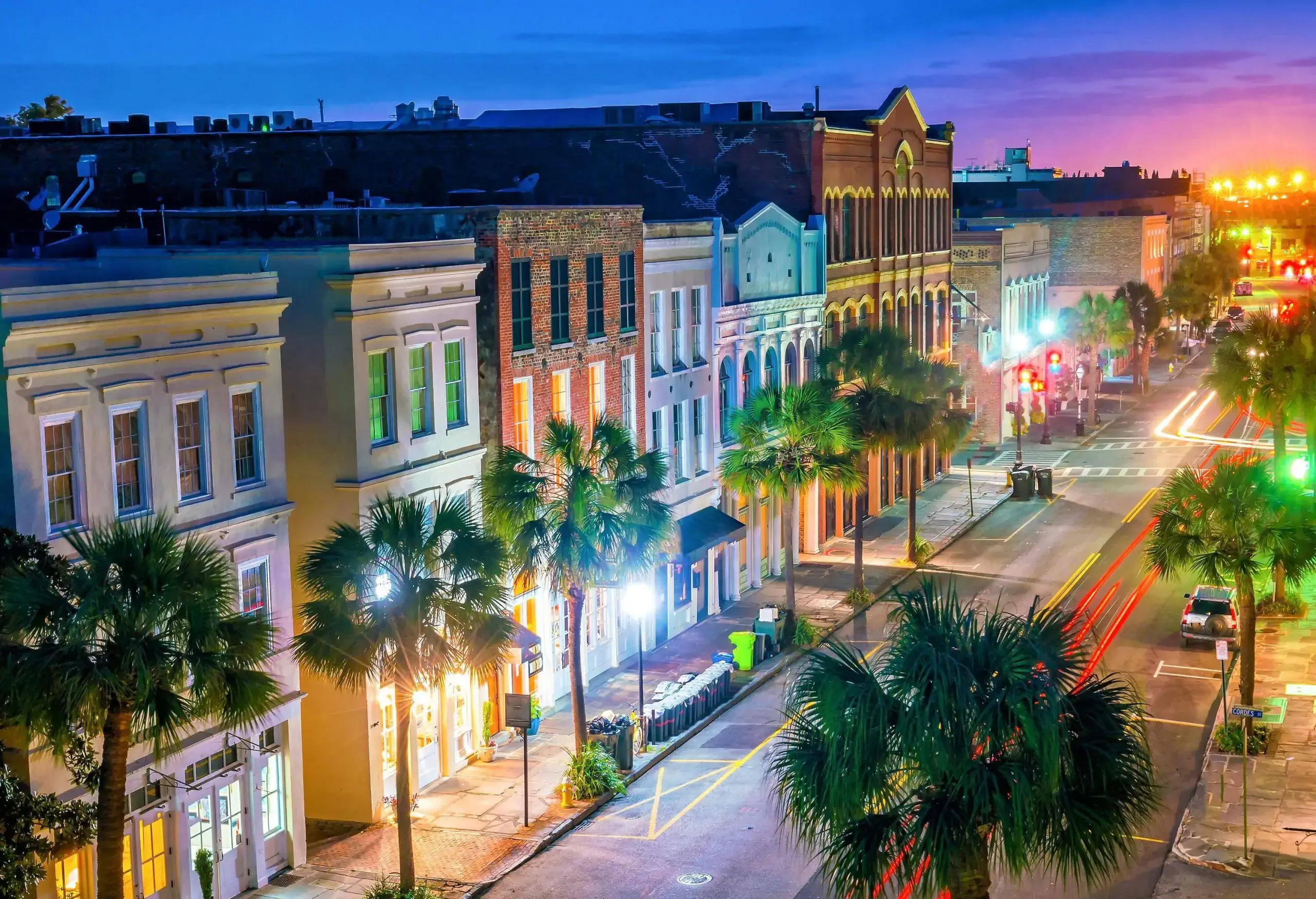 An empty street at night lined with trees and brightly lit lampposts amidst the colourful buildings.