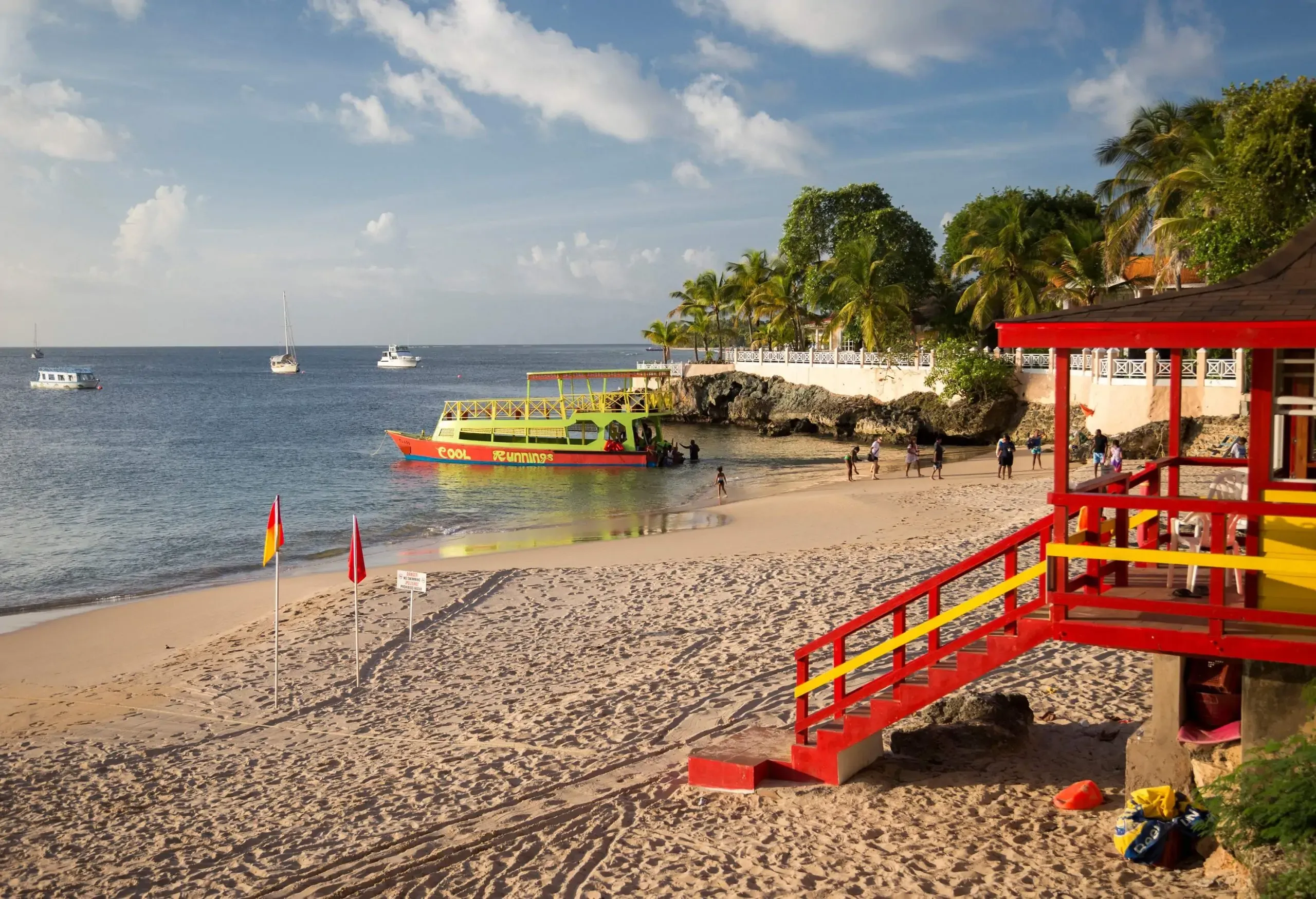 A red lifeguard tower overlooks a beach with tourists disembarking from a boat.