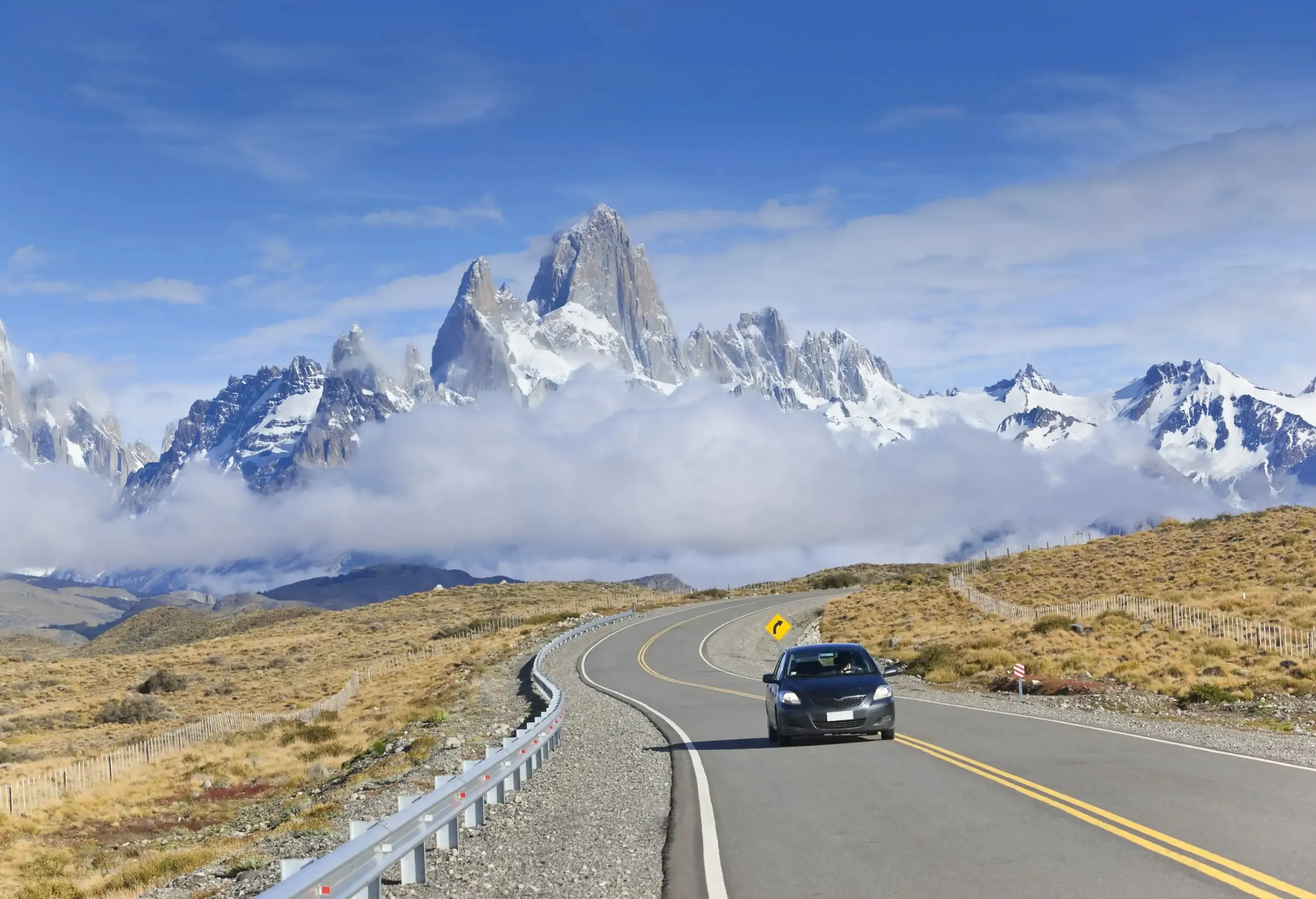 Rising triumphantly in the background, the iconic Mount Fitz Roy, a striking granite peak that towers over the Patagonian landscape, shrouded in mist, creates a dramatic and unforgettable scene as a car navigates the winding road away from this natural wonder.
