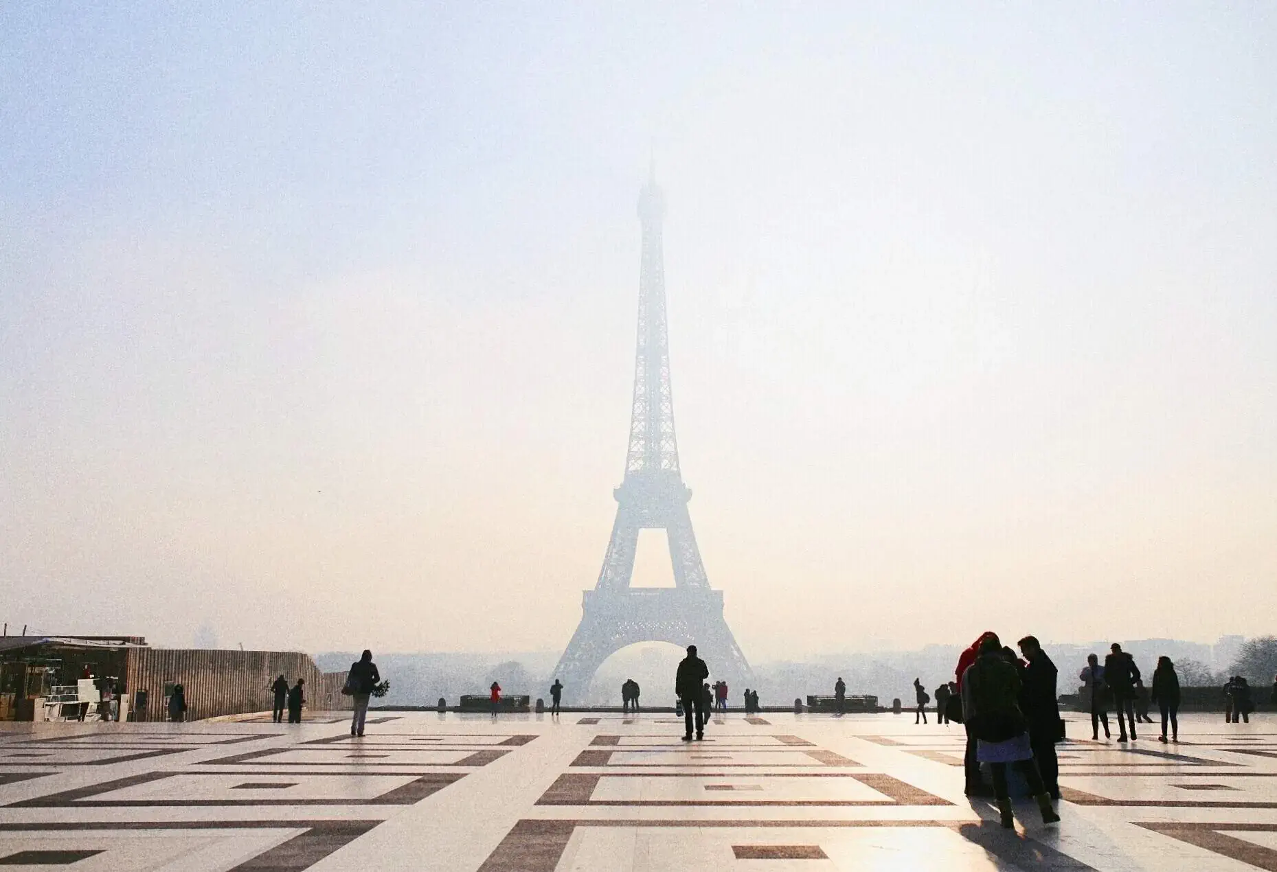 A plaza with a square tile design, where a large number of people admire the Eiffel Tower view obscured by a dense mist.