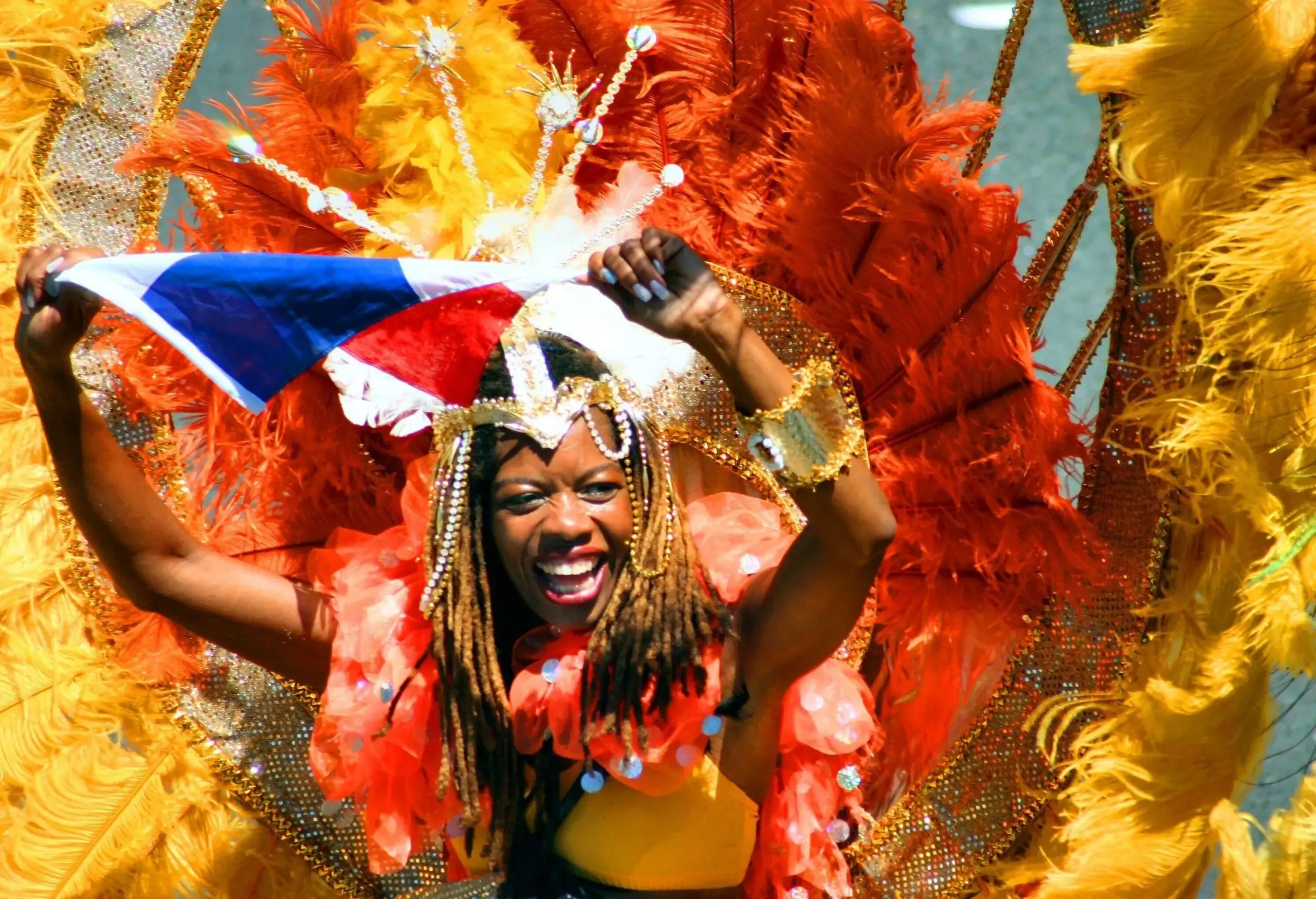 A dark-skinned woman wears a colourful feather backpack wing and a big smile while holding a piece of clothing during a festival.