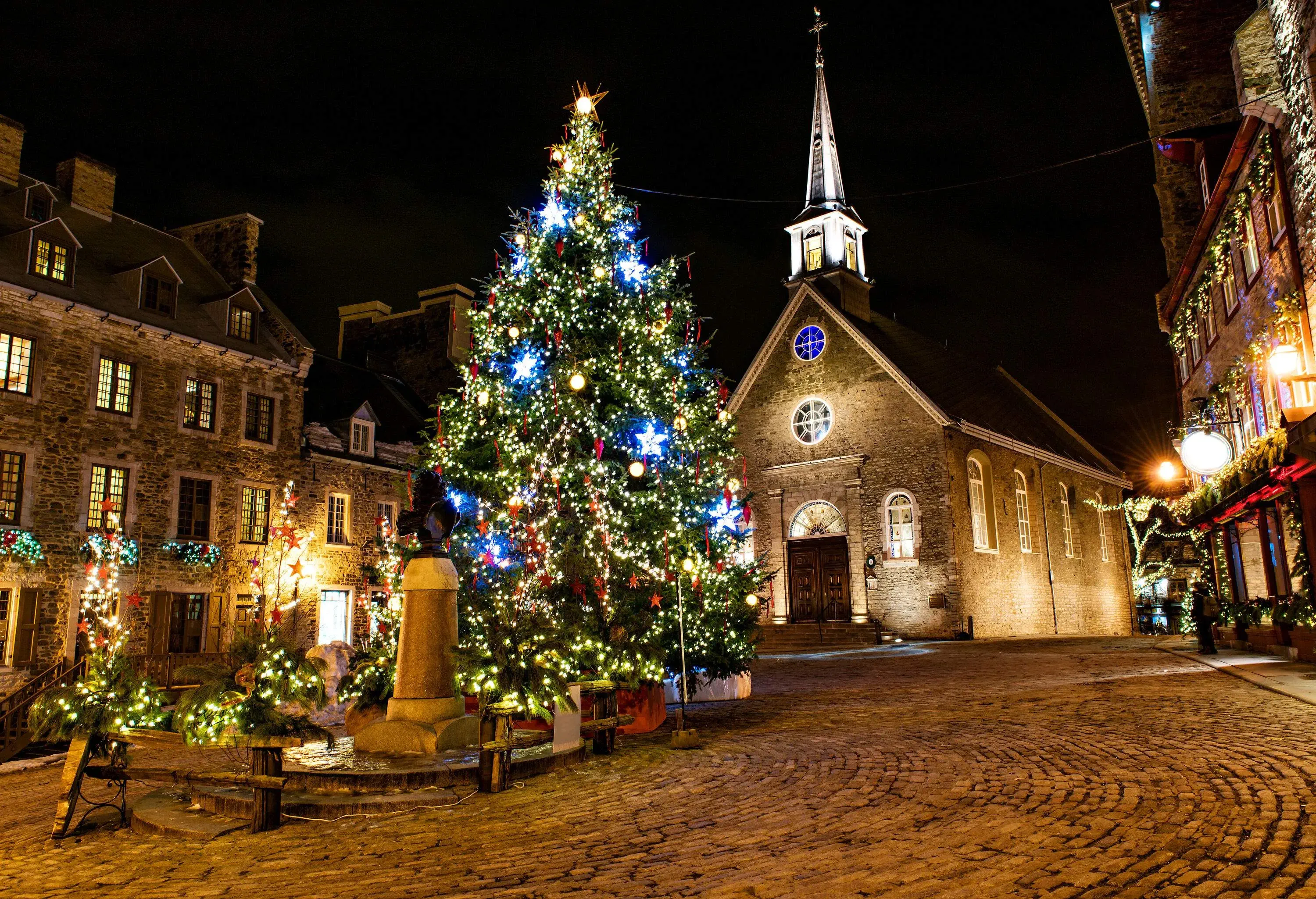 An illuminated Christmas tree in the centre of brightly lit houses in front of a tiny church.