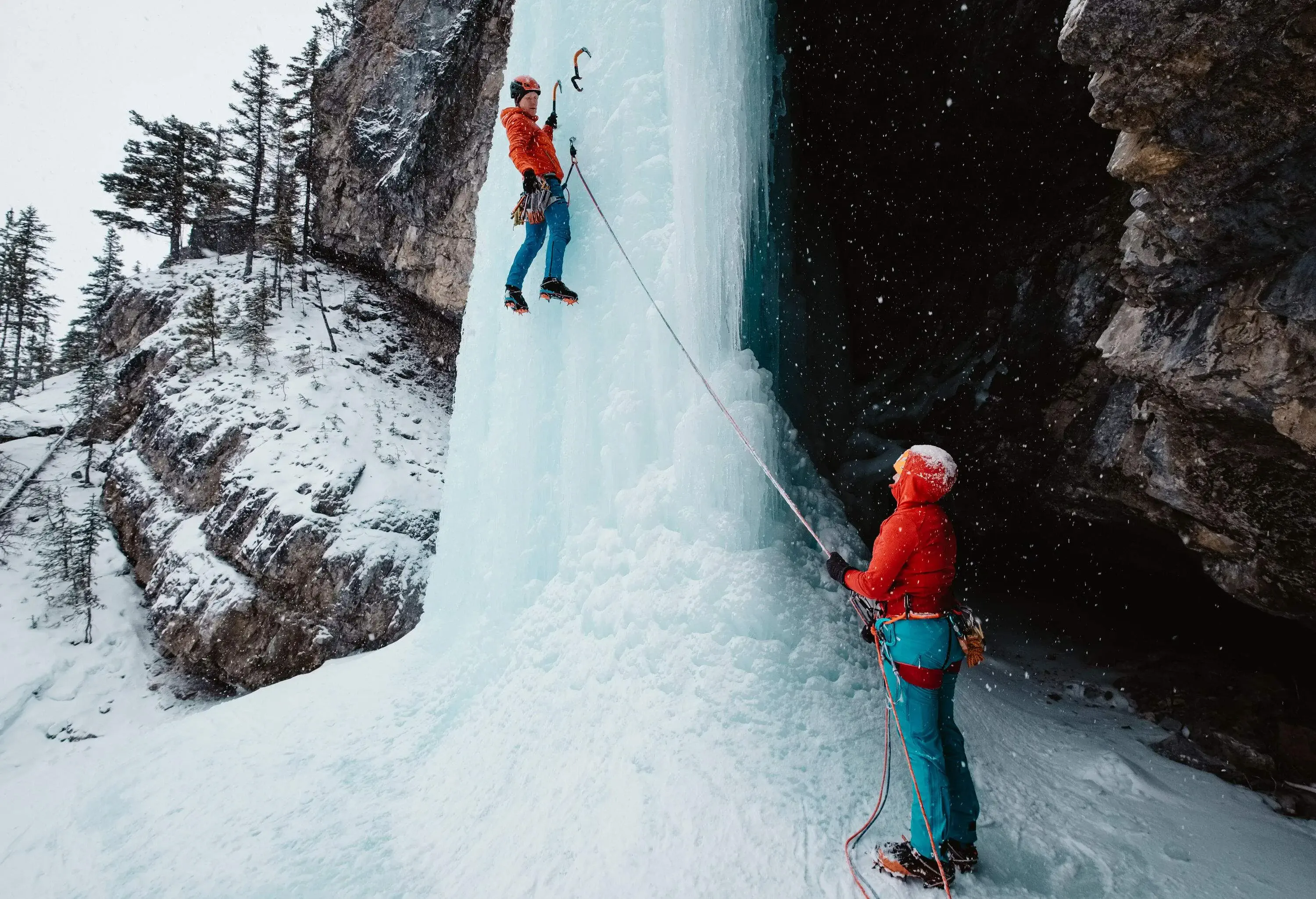 A male ice climber hangs on the steep frozen waterfalls with his ice axes firmly placed on the ice and looks down at his partner holding onto the rope in the foreground.