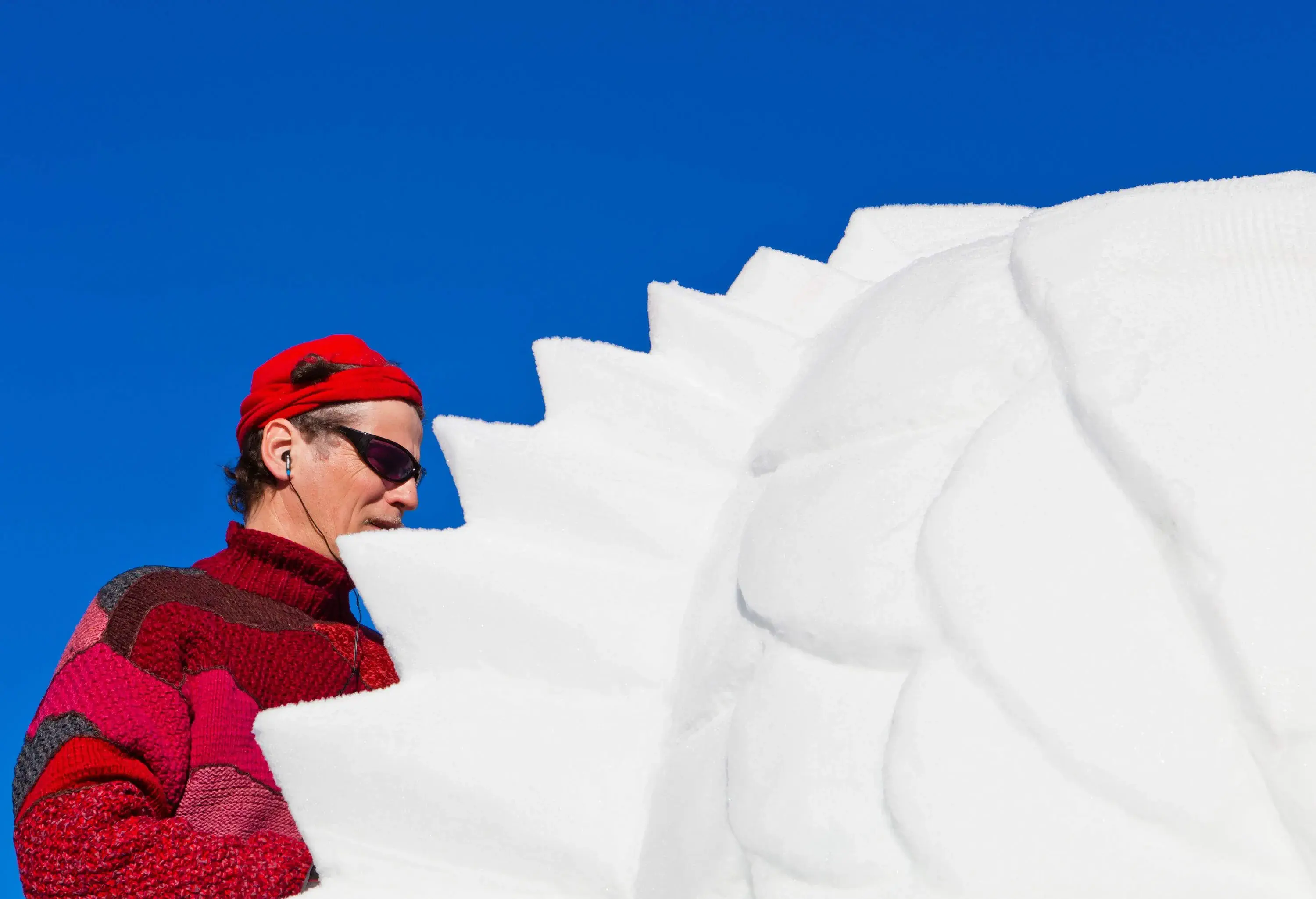 A man with sunglasses and wired earphones carving a beautiful snow sculpture outdoors.