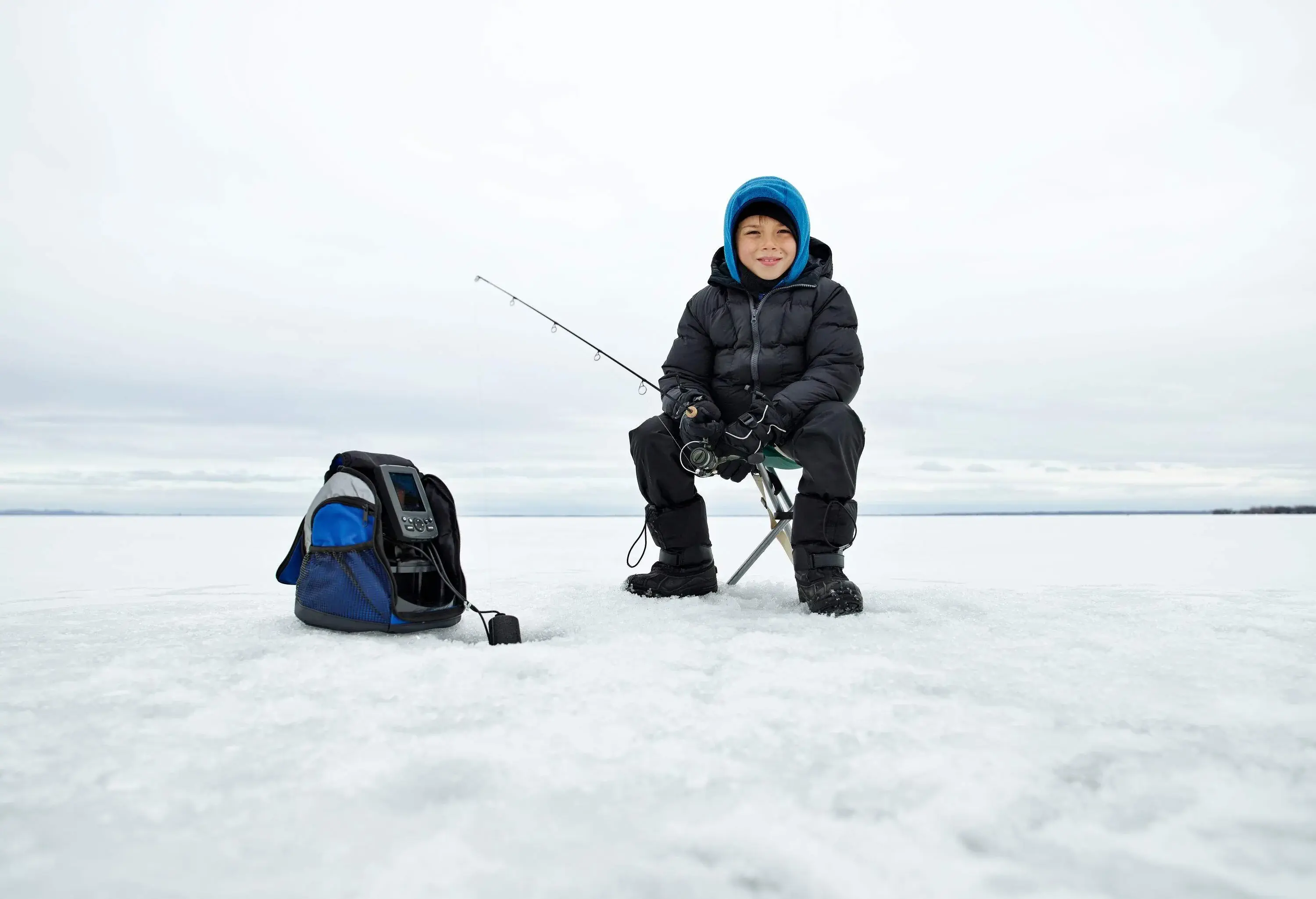 A boy kid sits on a folding chair and holds an ice fishing rod, patiently waiting for a catch using an electronic fish finder.