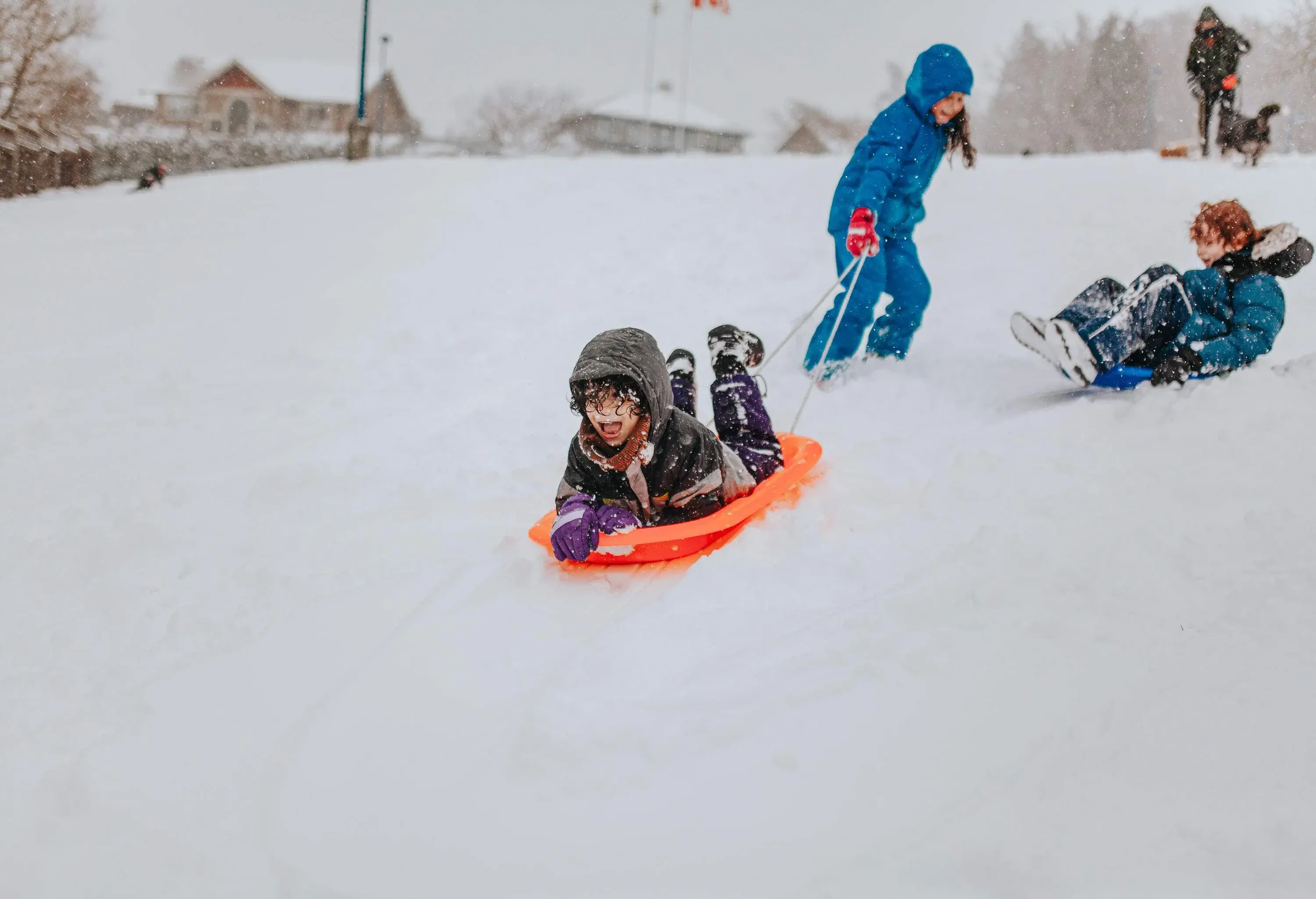 A kid in winter clothing lies on an orange sled being pulled by a child during snowfall.