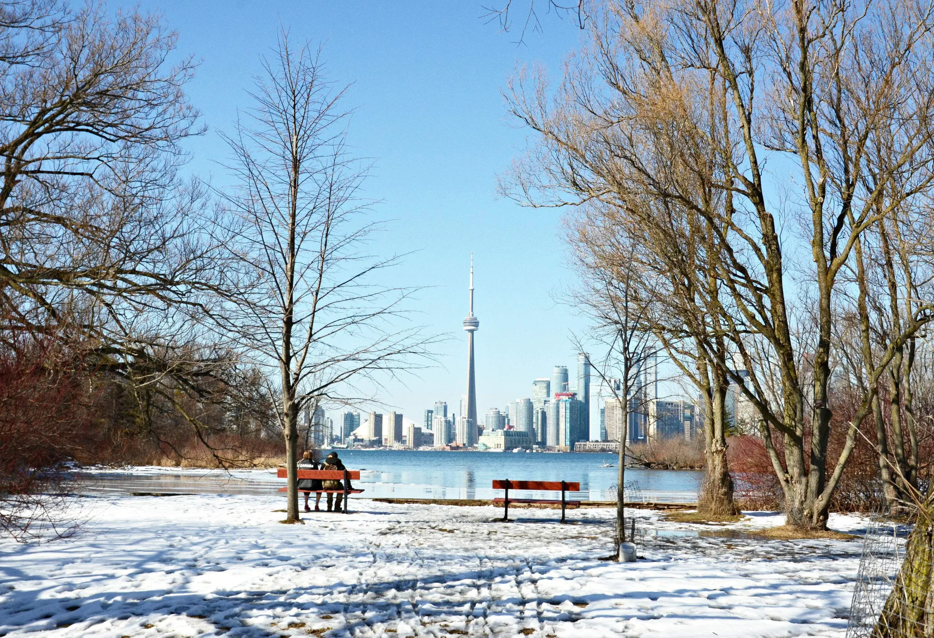 Two people sitting in a snowy park looking at the cityscape across the river.