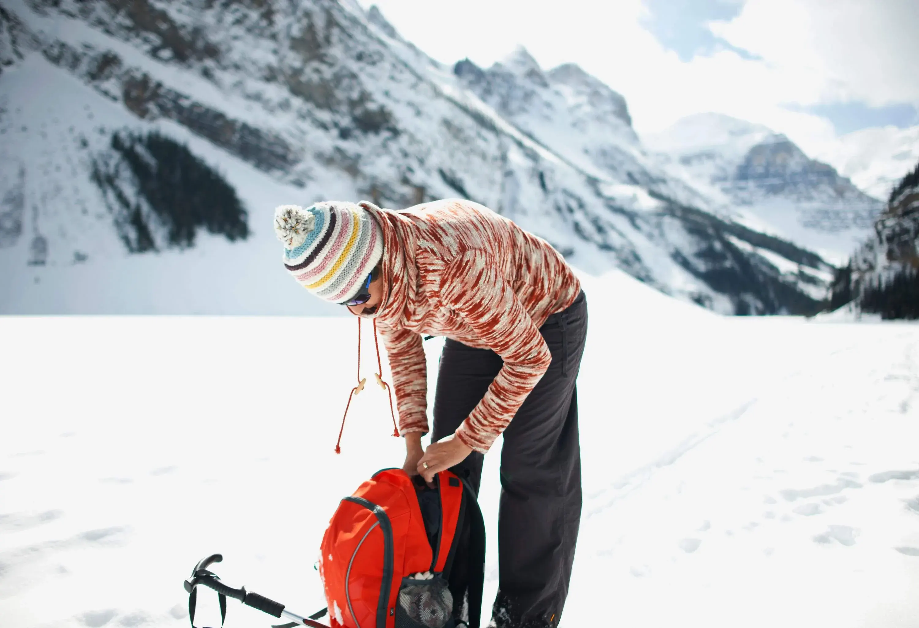 A person in colourful winter clothing bend over his red bag on the snow-covered land beneath the snowcapped rocky mountains.
