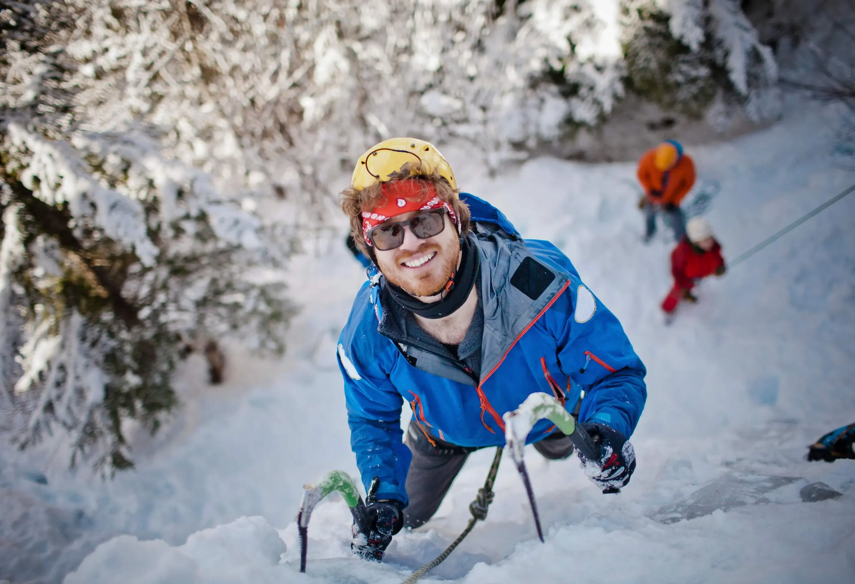 A man in a blue jacket, sunglasses, and orange helmet smiles as he climbs an ice cliff using a pair of ice hooks.