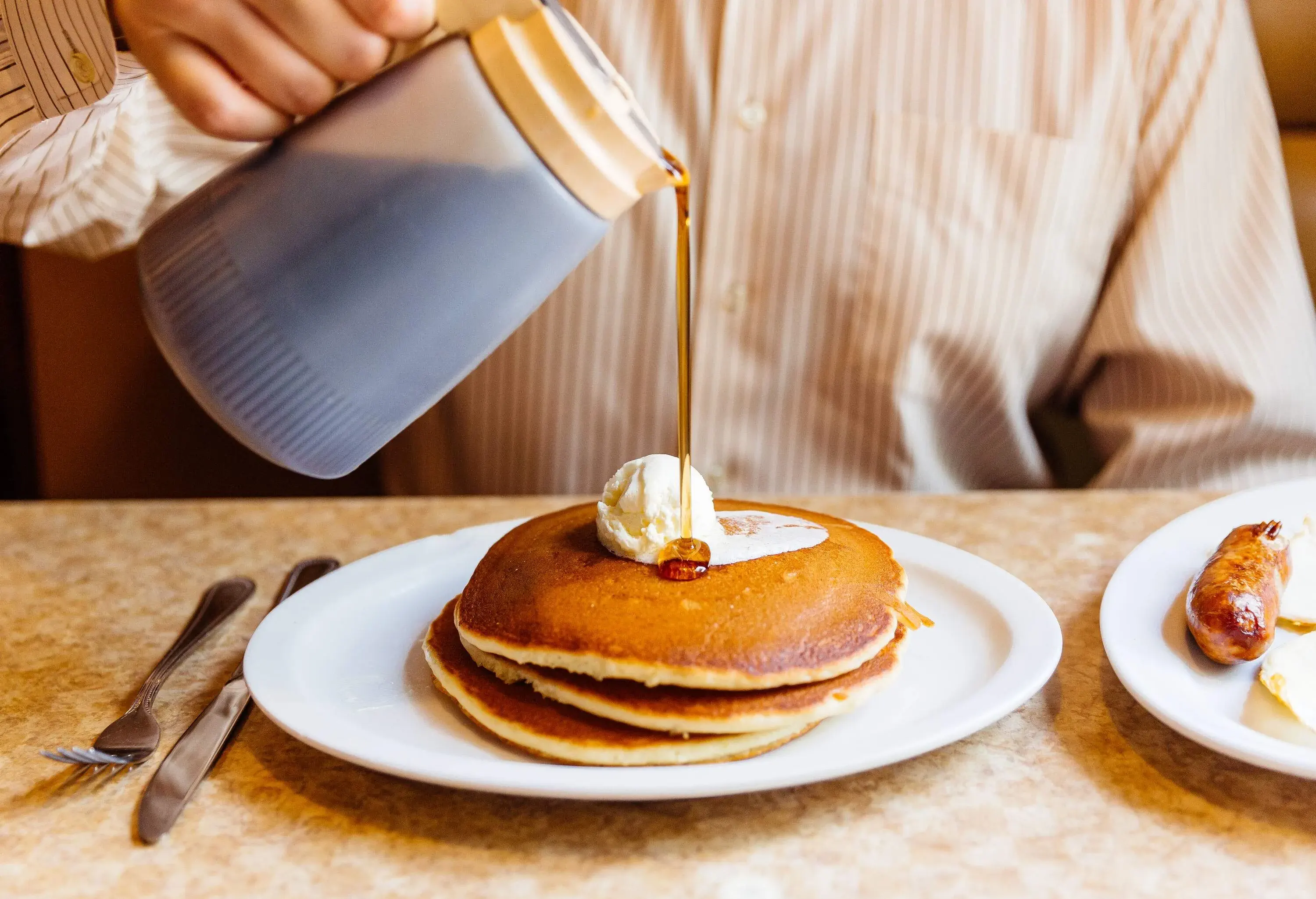 A stack of three fluffy pancakes is served on a plate, with a person pouring a generous amount of maple syrup over them while a scoop of butter melts on top.