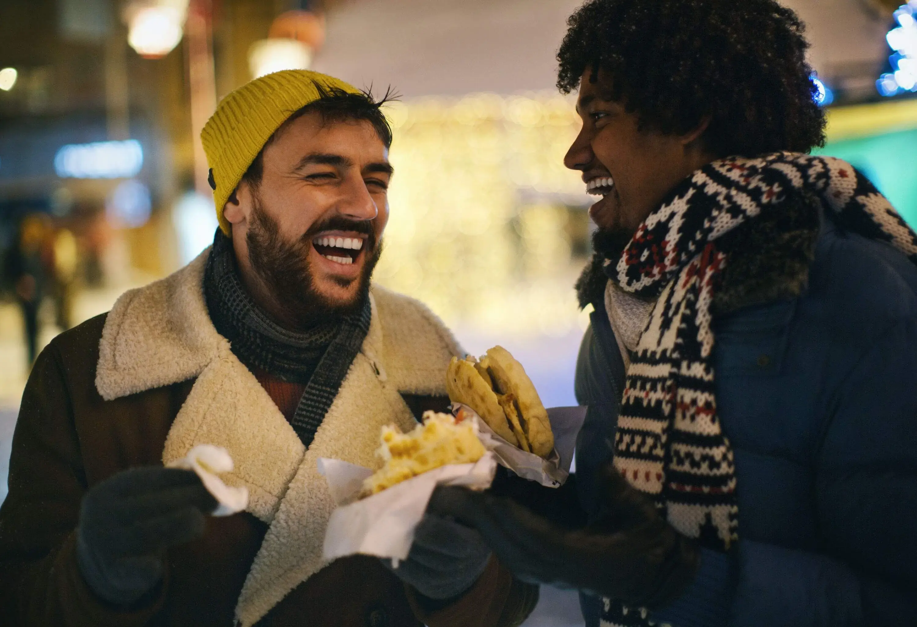 Two people dressed warmly laughing together while enjoying street food.