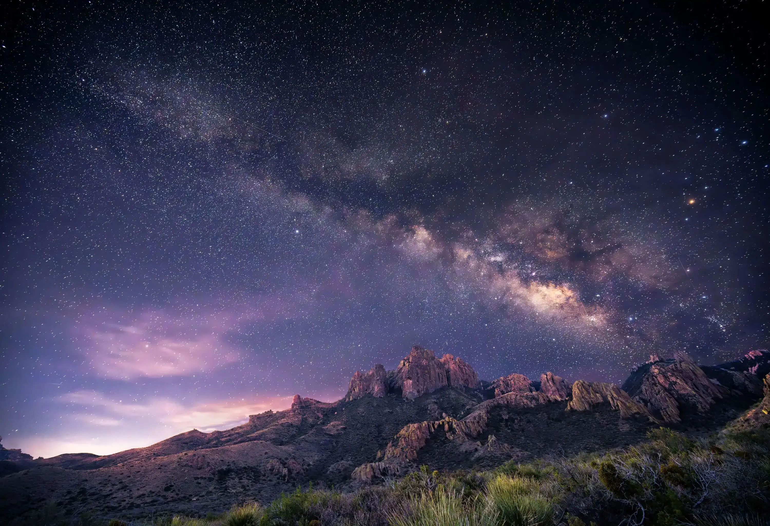 A star-filled night sky with the Milky Way stretching over a rugged mountain landscape at dusk.