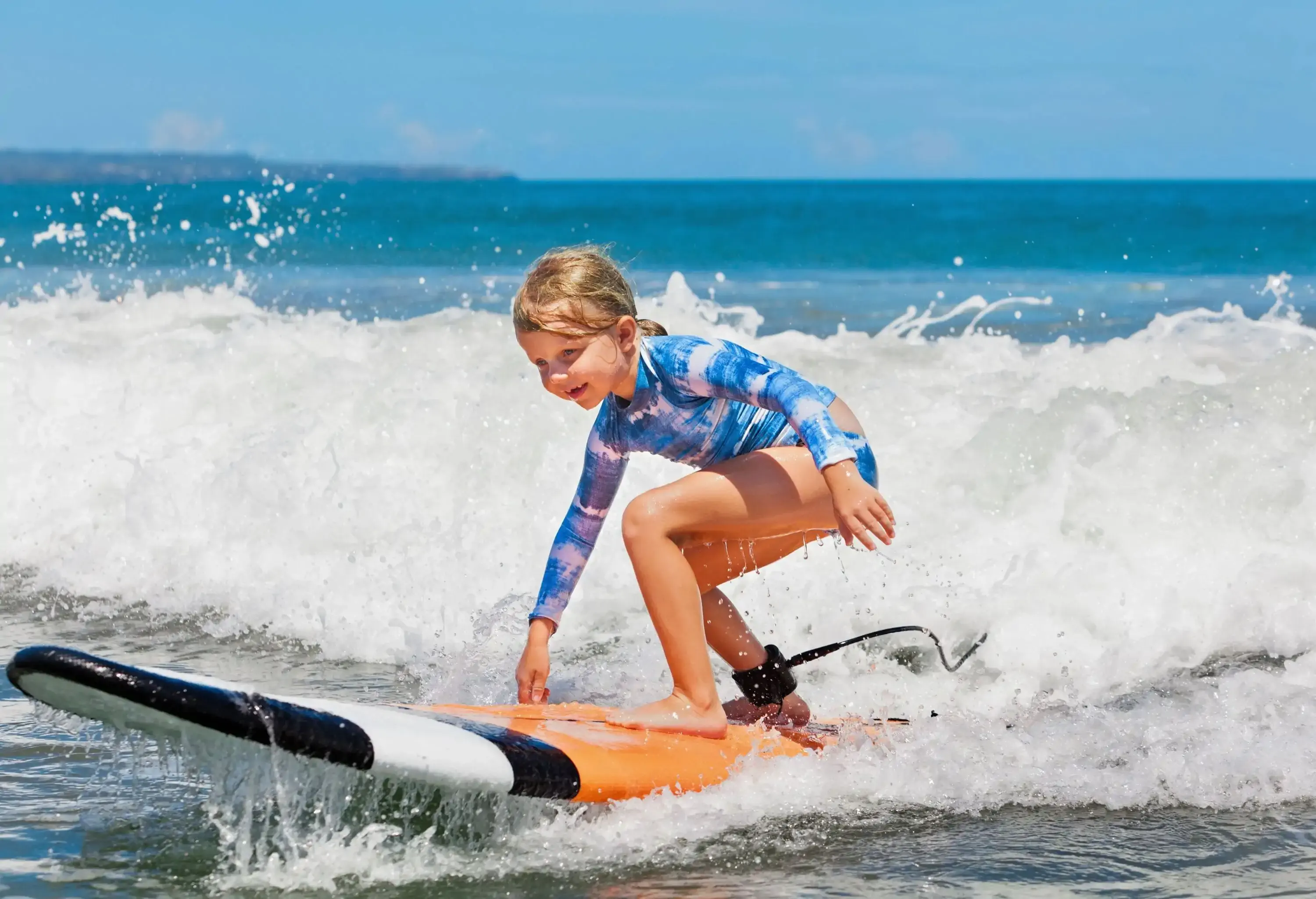 A young blonde girl smiles as she balances on a surfboard with crashing waves behind her.