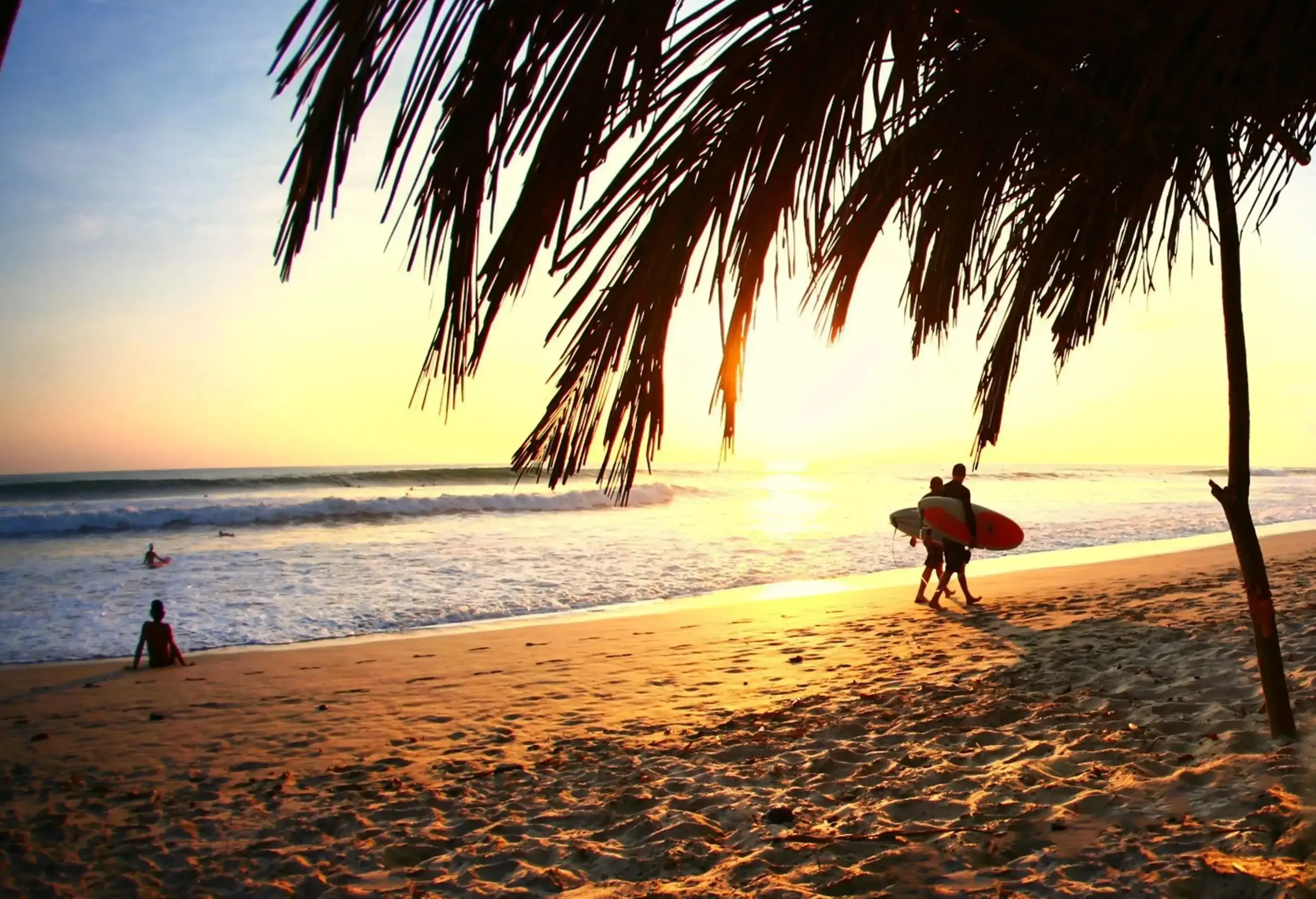 Two individuals strolling along the beach with surfboards on their sides, the sunshine shining off the water.
