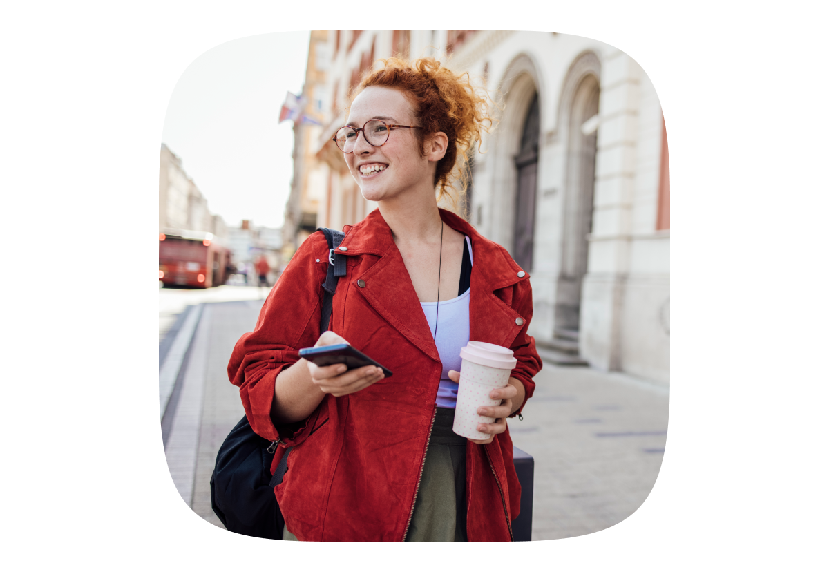 woman with coffee and phone in hand smiling and walking down the street