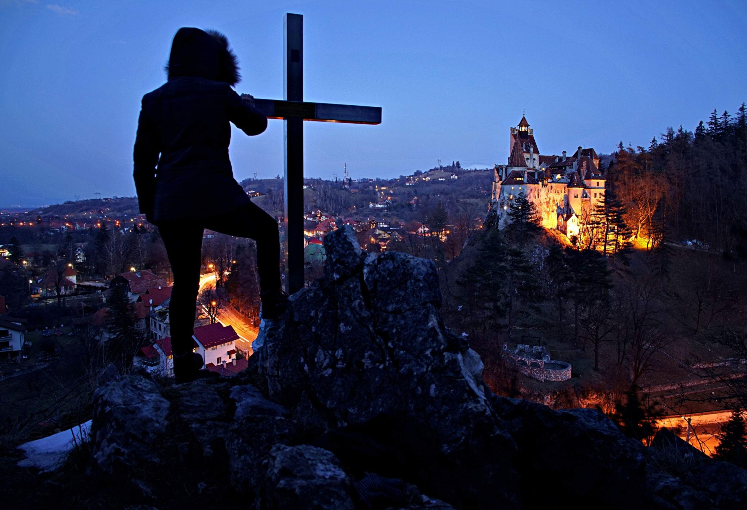 A silhouette of a person standing next to a cross embedded in a rock, with a distant view of an illuminated castle.