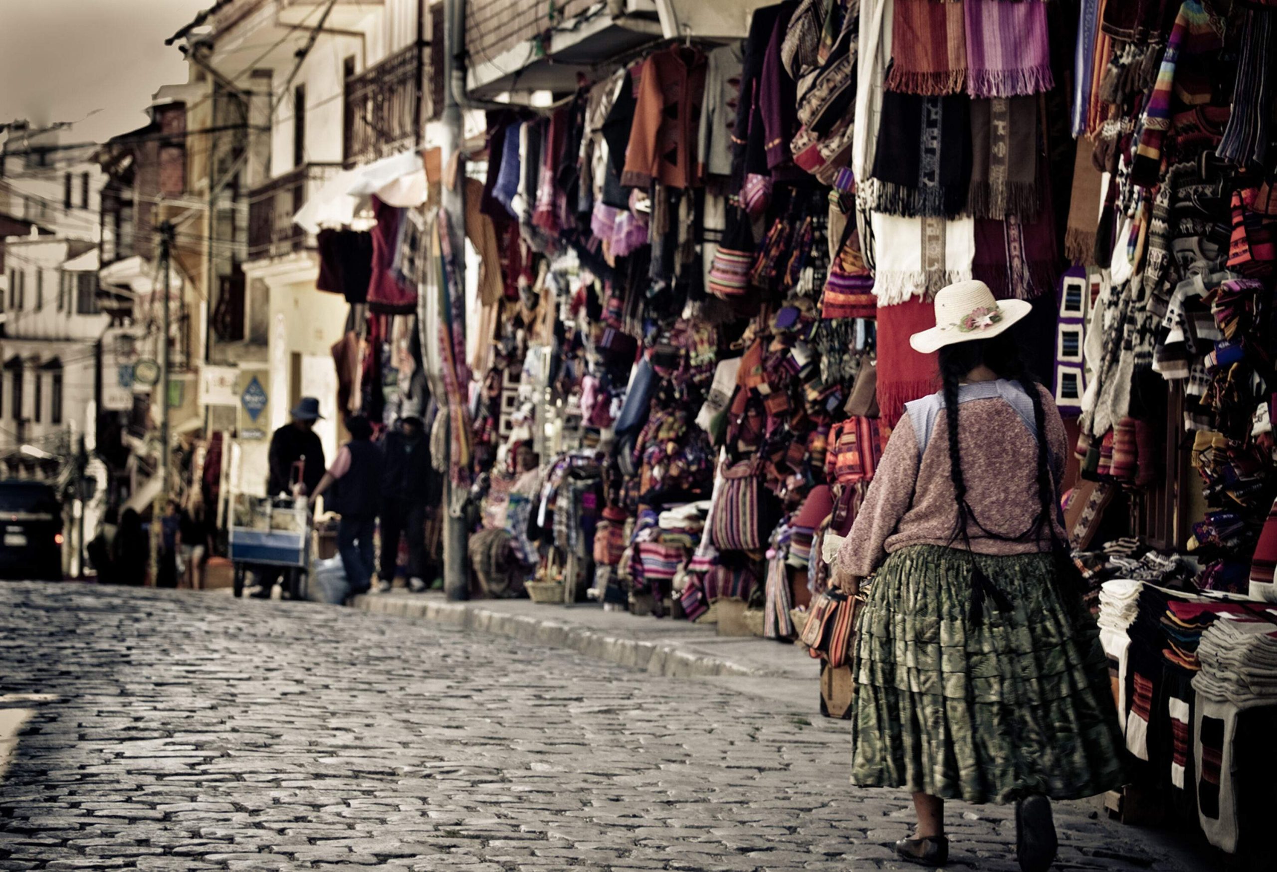 A woman in a skirt and hat walks on a cobblestone street along vendor stalls selling a variety of souvenirs.