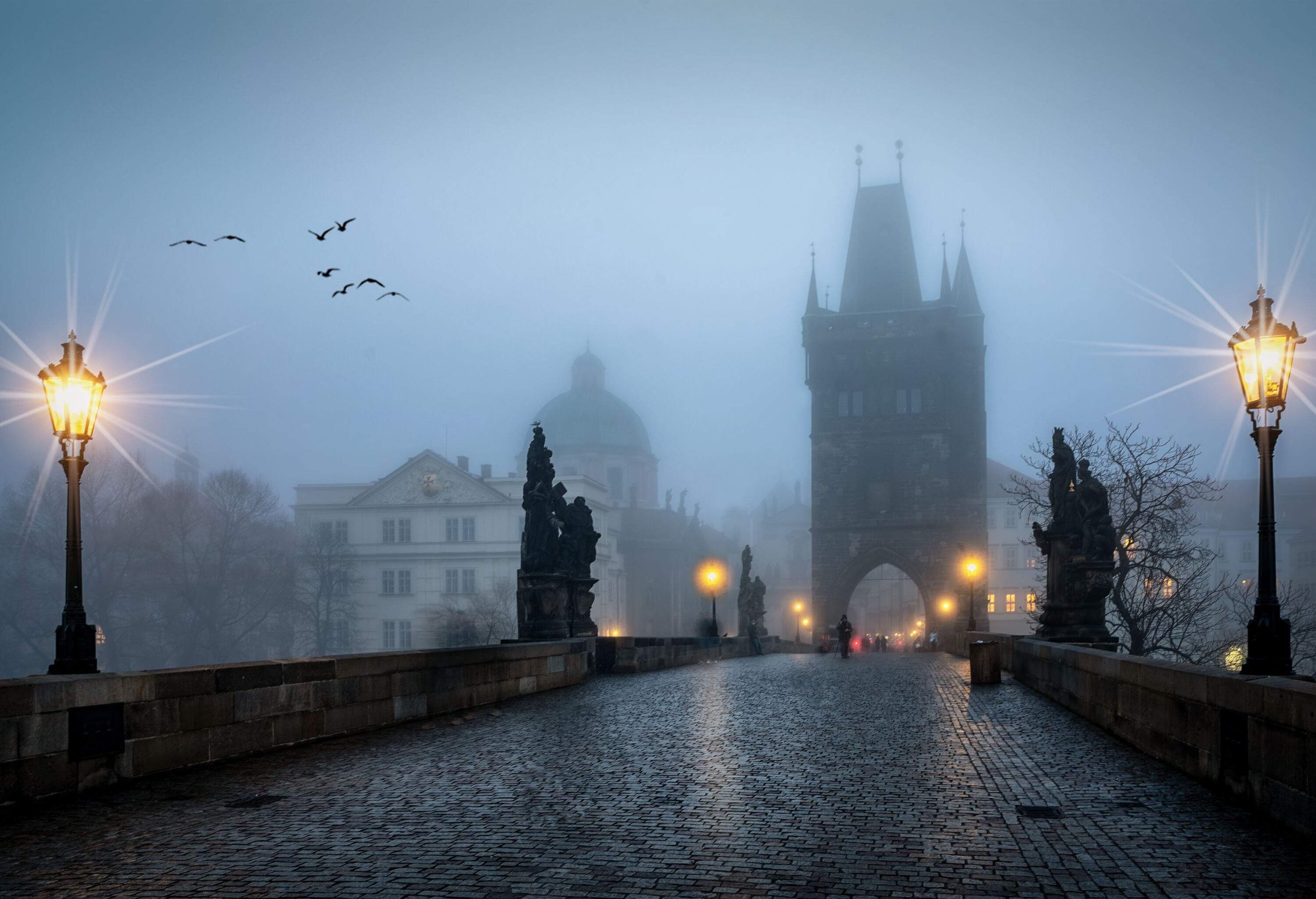 A medieval stone bridge with statues towards a castle and the city's old town on a foggy winter morning.