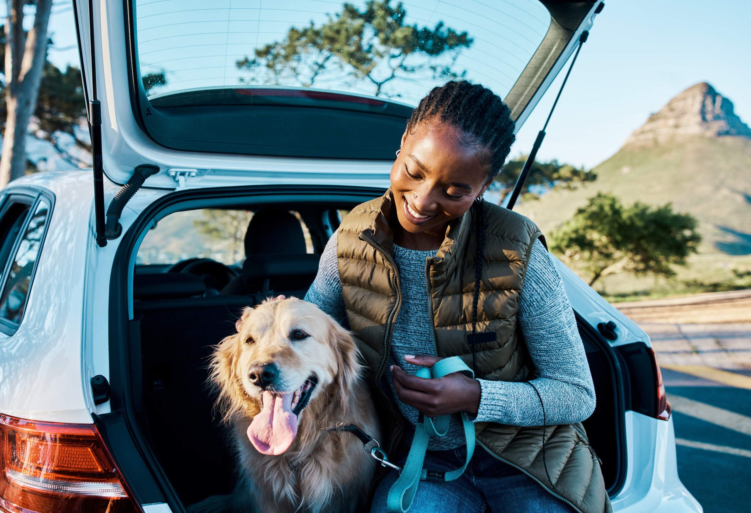 A smiling woman sits on the trunk of a car and pets her dog.