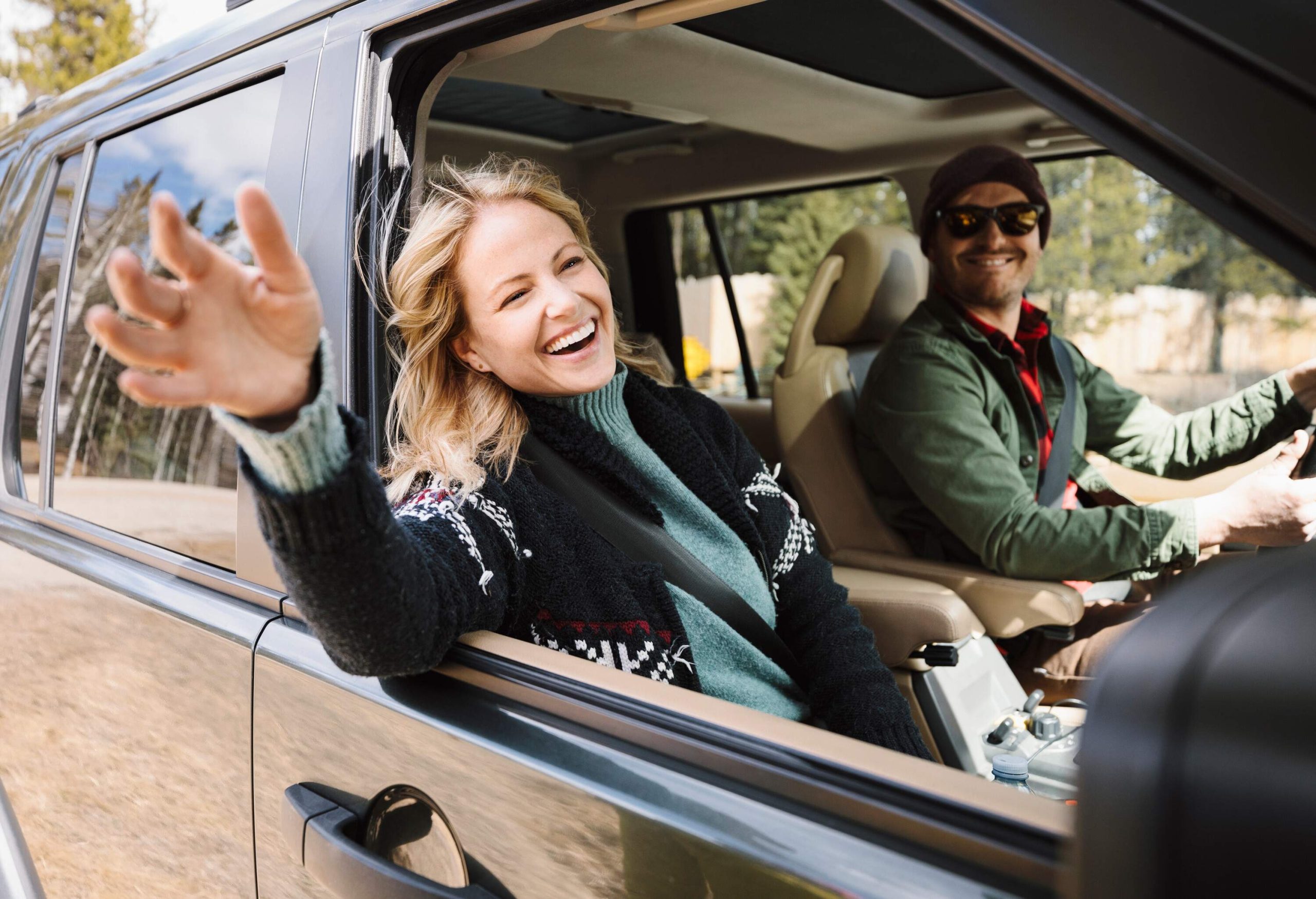 A couple sitting inside a car, with the woman waving her hand out the window and the man driving.