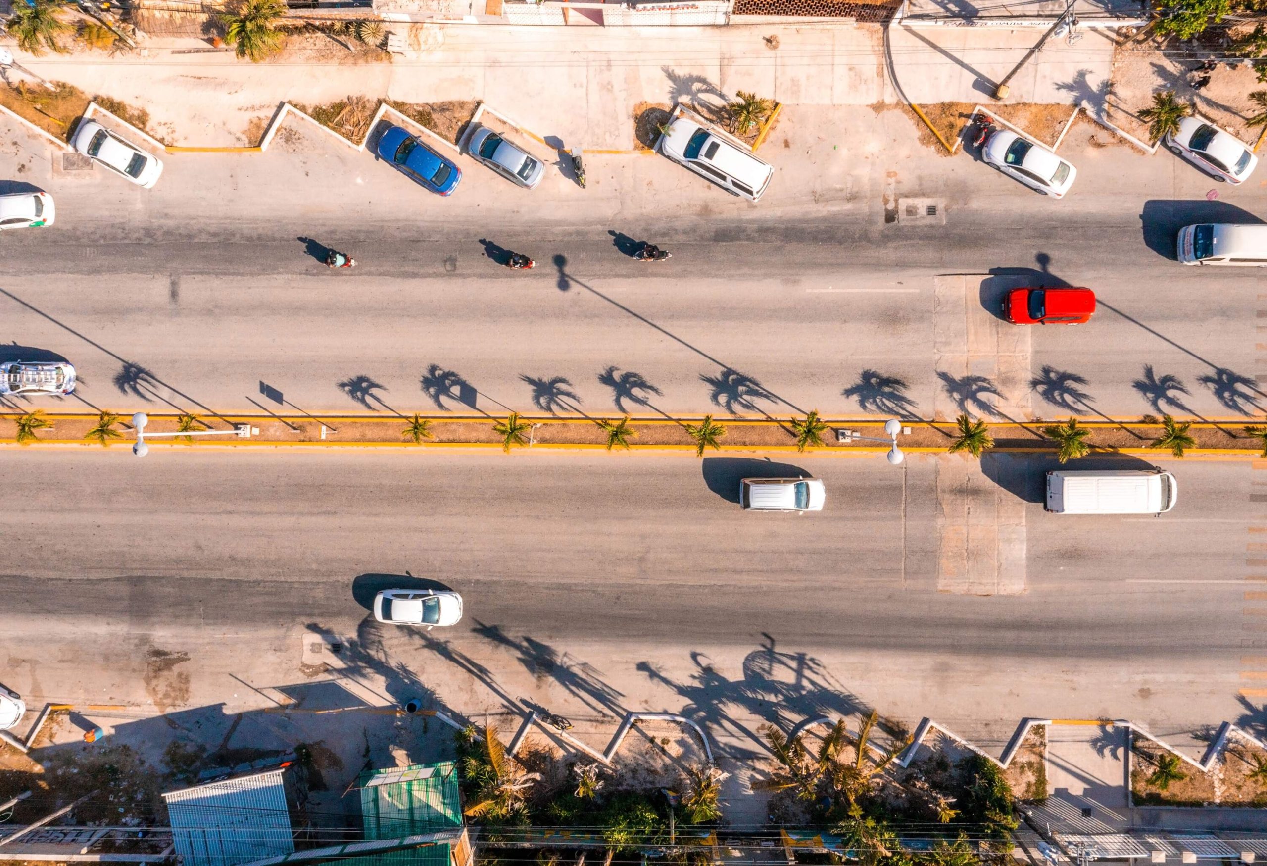 Cars travel on a two-lane highway lined with palm trees and parked cars.
